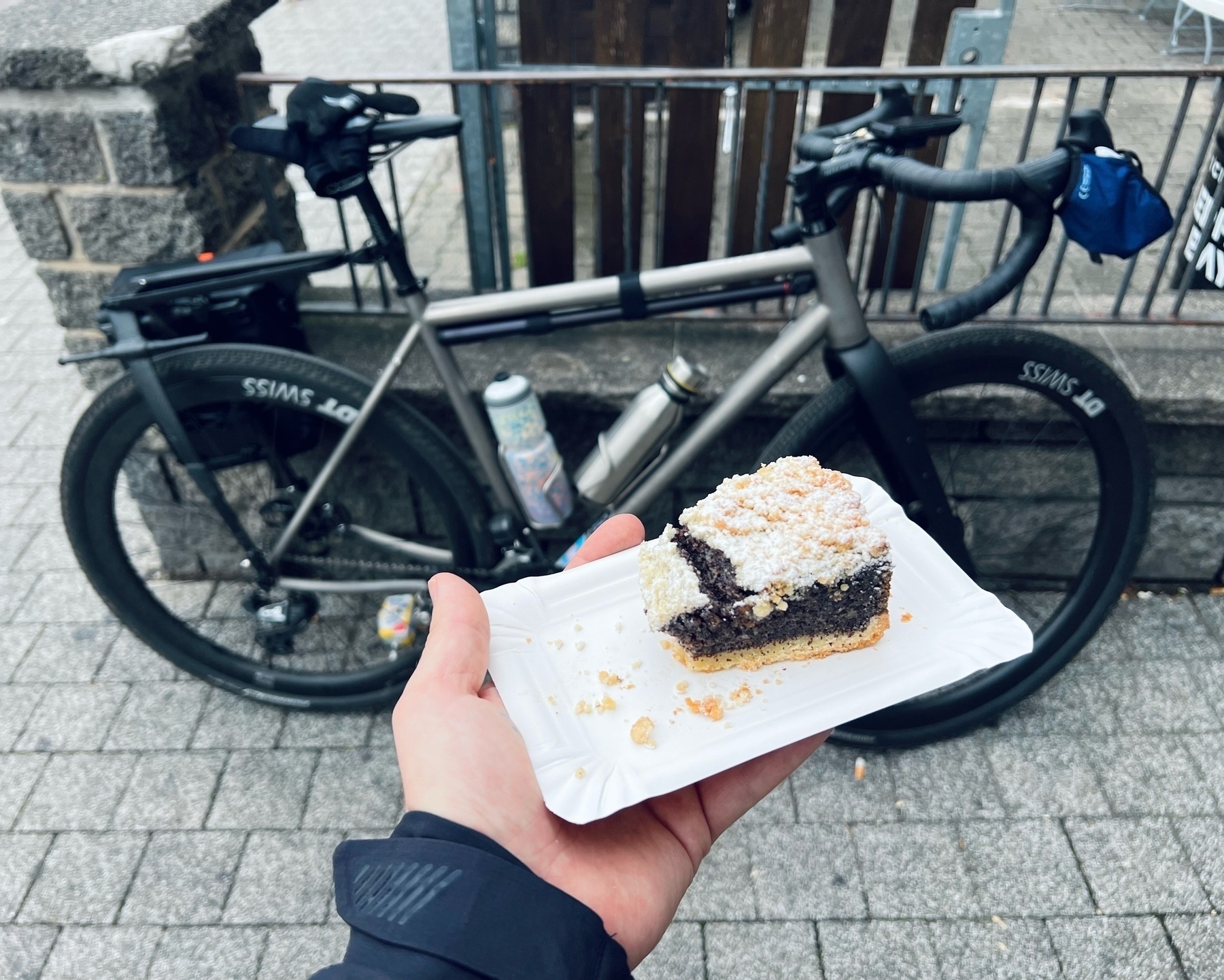 A piece of poppy seed cake ona rectangular paper plat held in the left hand of the photographer. in the background and slightly out of focus, a bike leans against a wall and fence. 