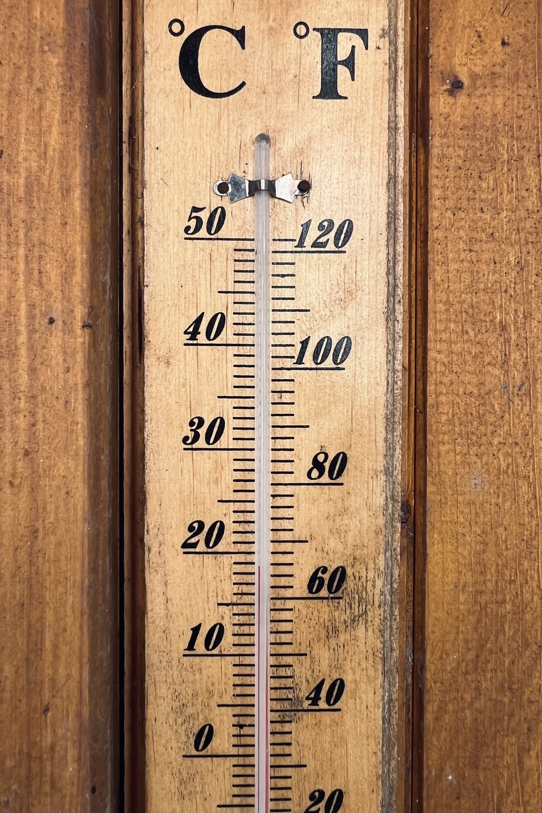 Outside thermometer with a wooden backing plate attached to a wooden wall of a garden shed and black lettering (Celsius on the left, Fahrenheit on the right). The mercury shows 19 °C.