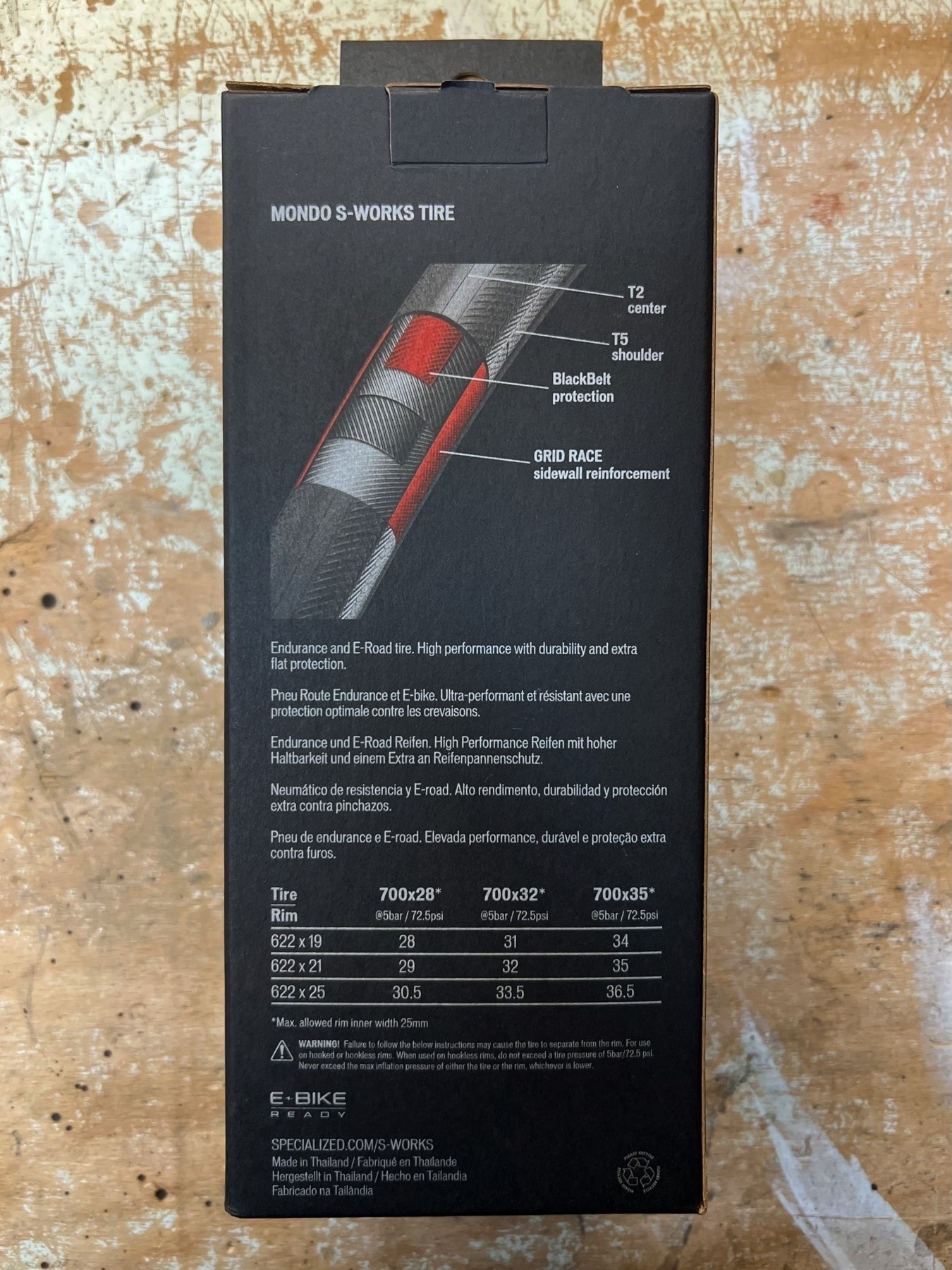 Back side of the black carton packaging of a bicycle tires laying on a wooden work bench. The packaging shows a cut-away diagram of the tire as well as other information. 