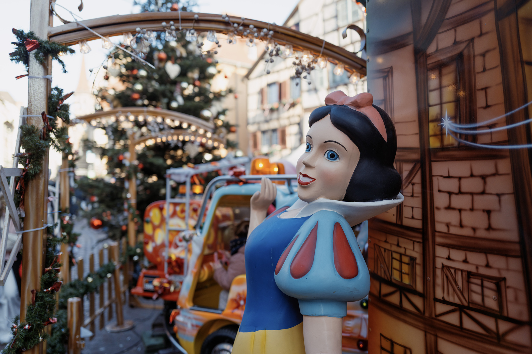 Life-size plastic figure of Disney‘s Snow White standing in front of the side of a Christmas market/fair ride. The right of the frame is dominated by a faux brown brick house. In the center background of the frame a Christmas tree is visible and various other decorations.