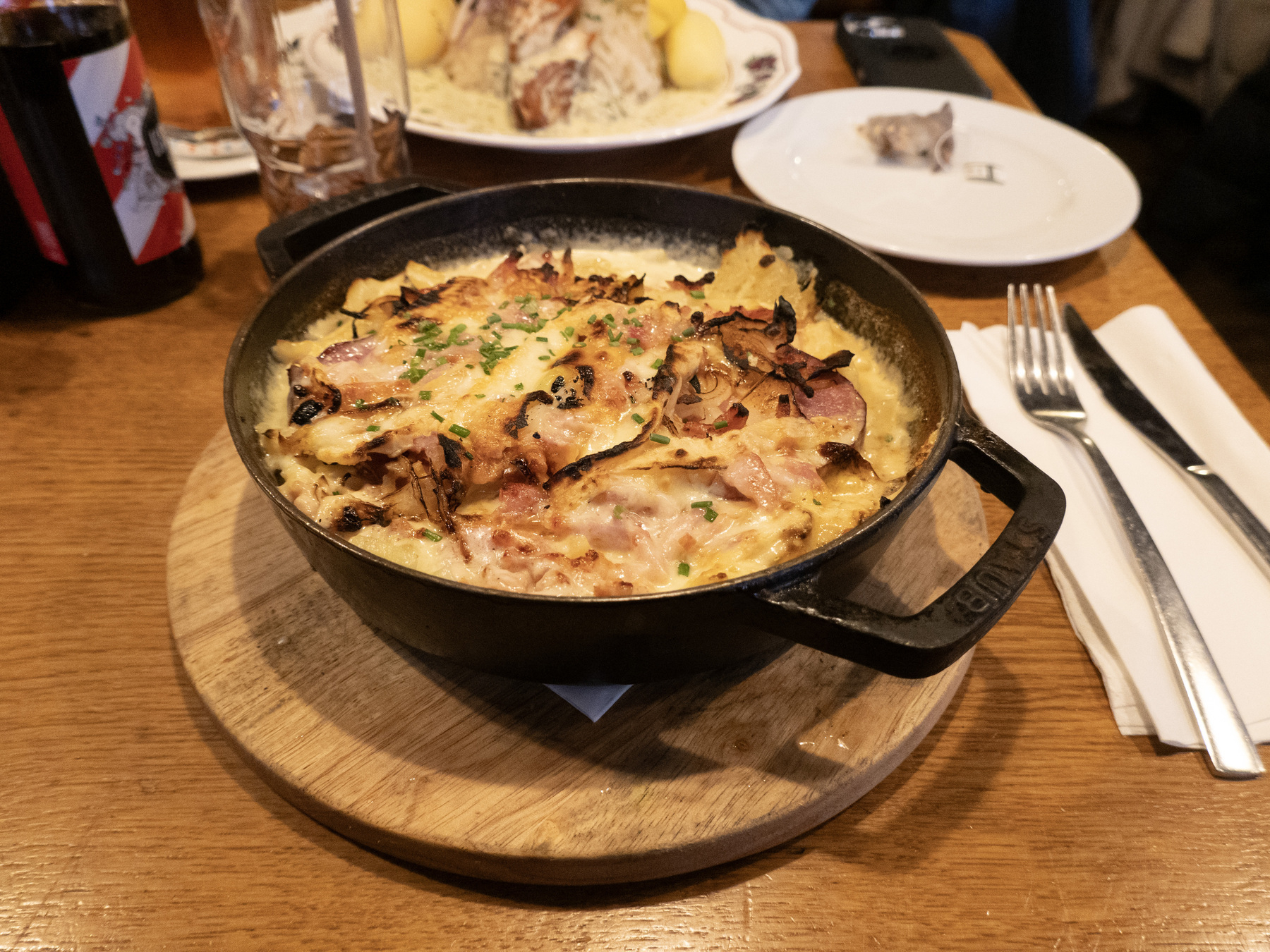 Alsac-style „Rösti“ in a cast-iron skillet on a wooden plate. To the right of the skillet a knife and fork lay on a paper napkin. Behind the skillet and away from the photographer the dish of a different diner is visible.