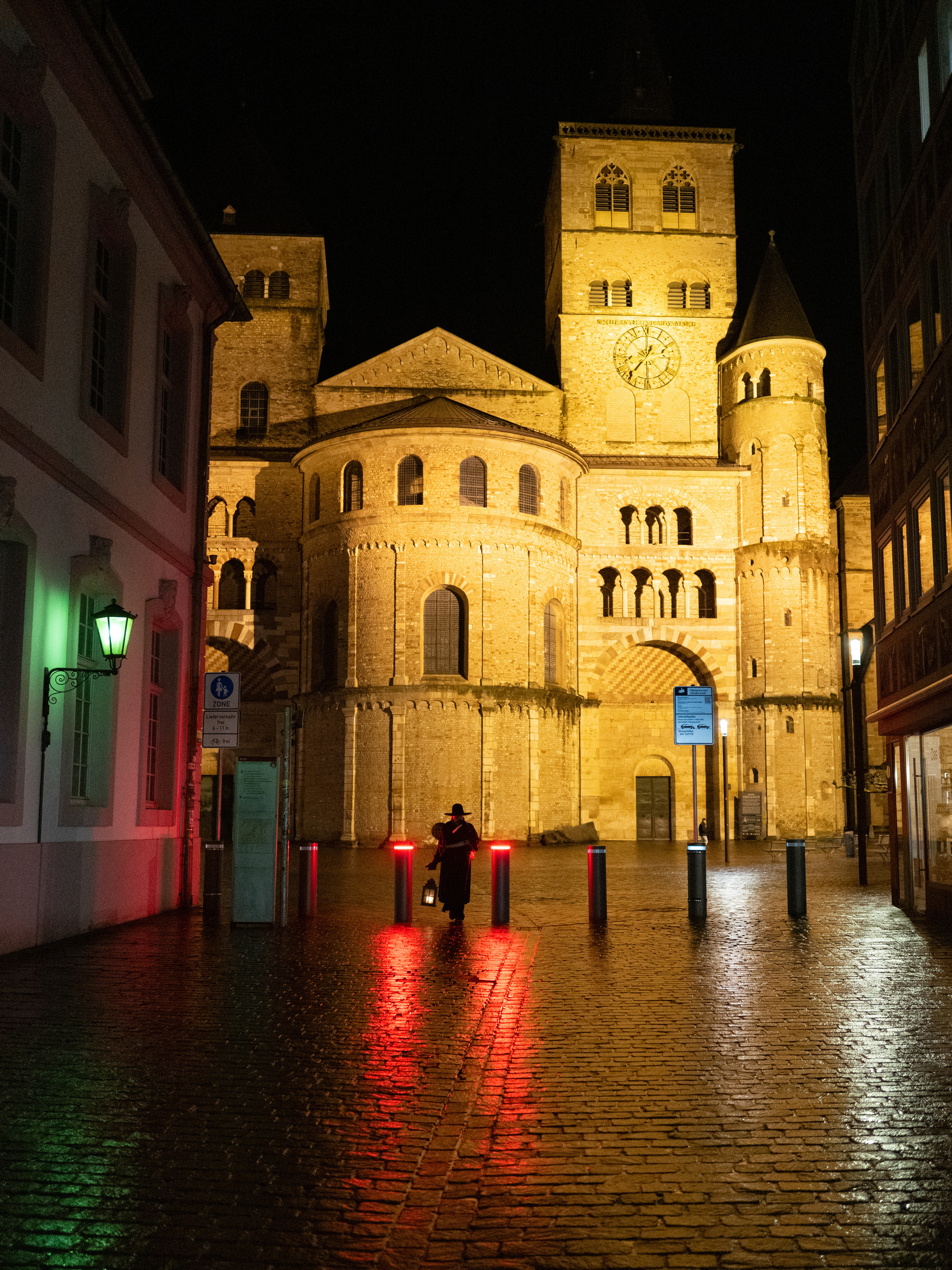 Night time scene. The cathedral of Trier and the square in front of it is visible in the background. The cathedral is lit from the outside. In the middle distance there are safety pollards separating the cathedral square and the passageway the observer is standing in. The pollards have a red light at the top and are illuminating the wet floor in front of them. Between two of the pollards a dark figure clad in a long trench coat and a wide-brimmed hat approaches. They are carrying and old lantern. 