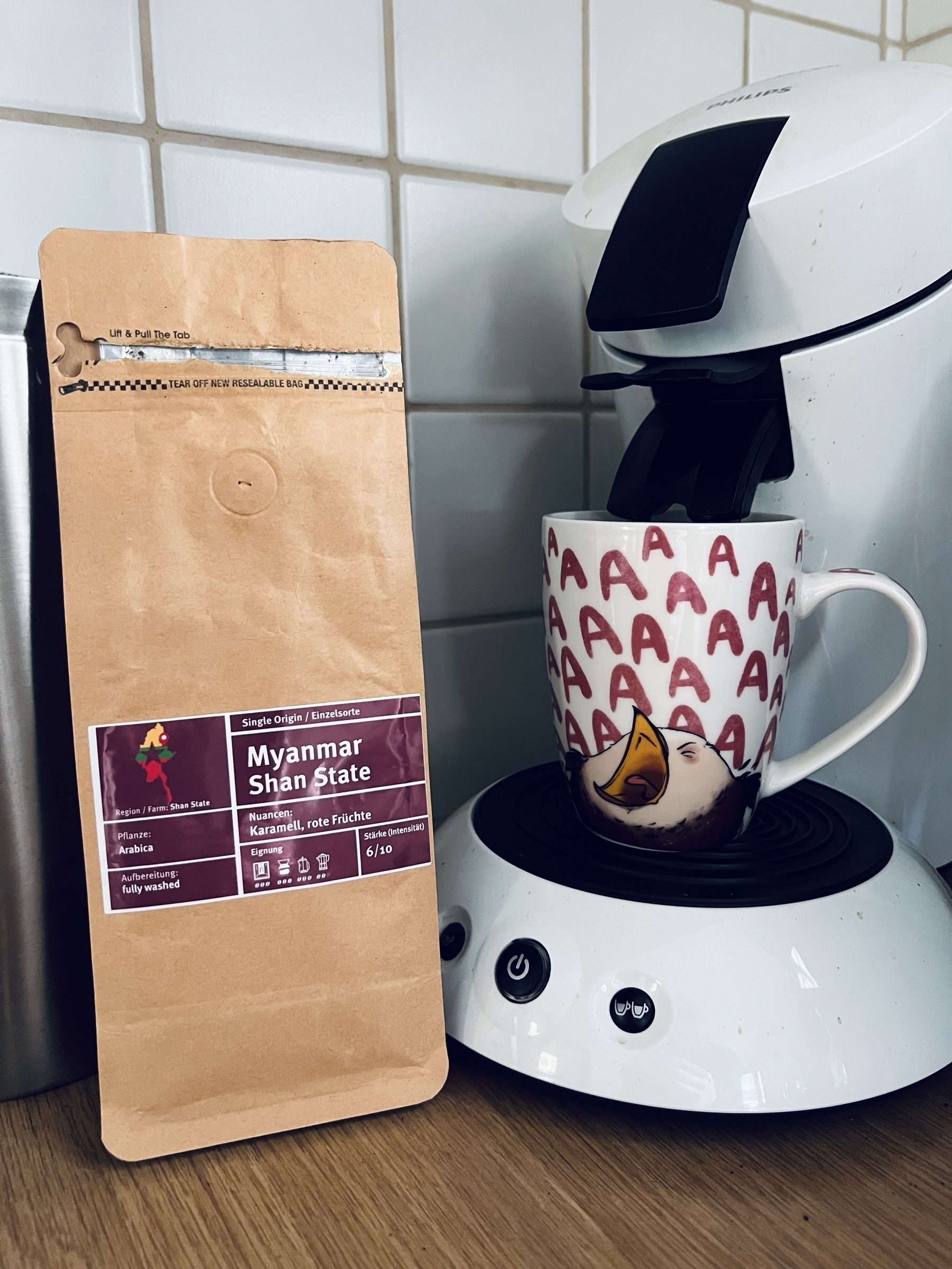 Small coffee packet standing next to a Philips Senseo coffee maker. The coffee maker has a mug on it showing a bird screaming in frustration surrounded by large letters A.