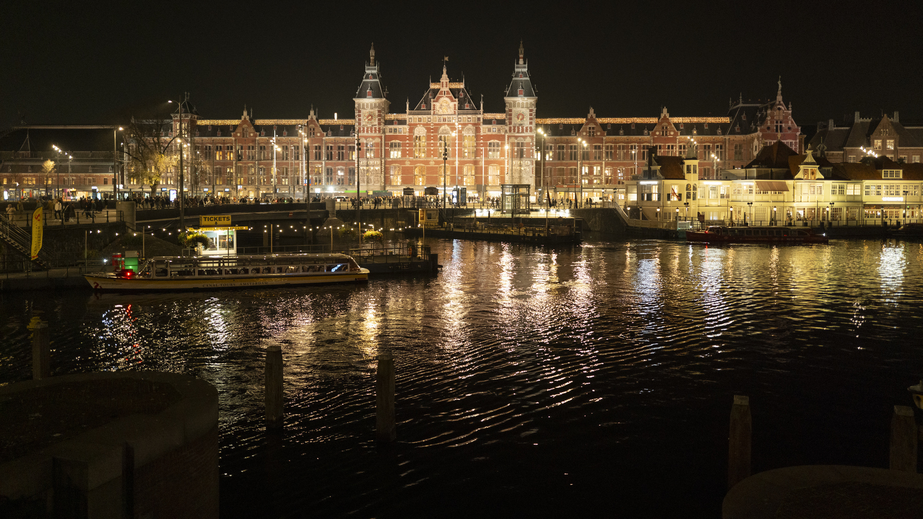 Nighttime shot of the Amsterdam Centraal station taken from across the canal right in front of the station. The lights appear in a warm yellow and orange and illuminate the square in front of the station as well as the water further towards the photographer.