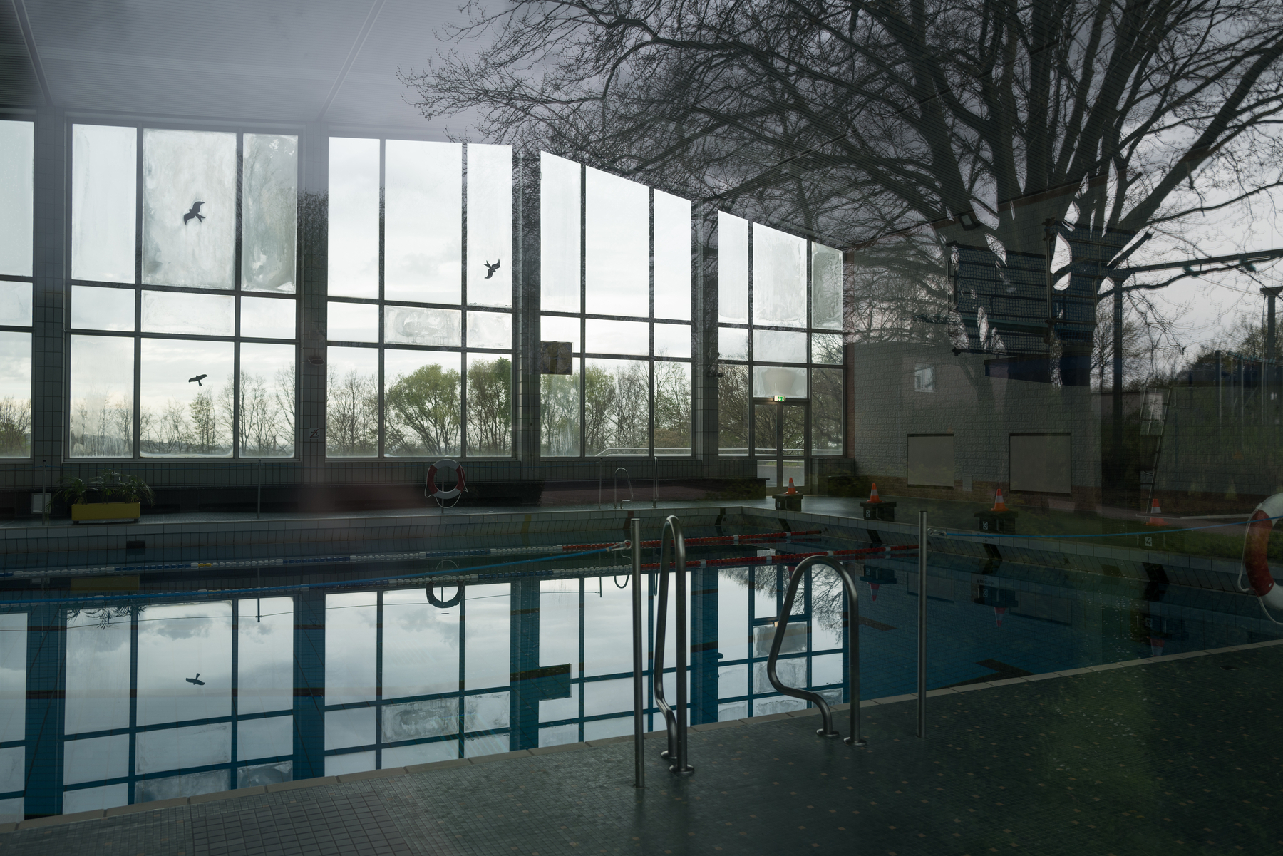 Photo of a large indoor swimming pool shot through a window looking in at an angle. The pool is completely deserted and in the windows reflections of the water, the sky, and a tree that is behind the photographer can be seen. 