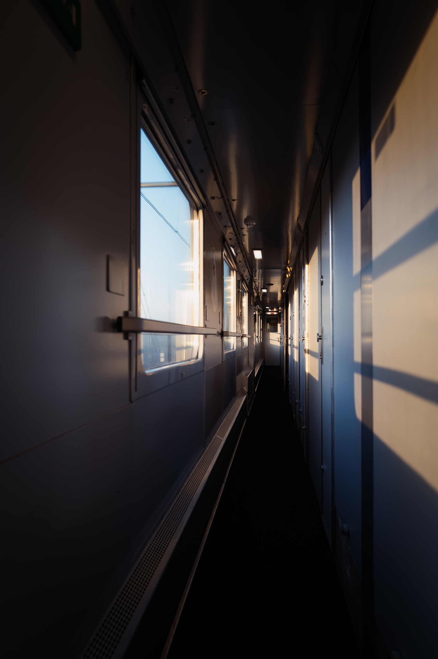 Photograph of the hallway of a train sleeper car. All doors to the compartments are closed and light is streaming in from the left-hand windows, casting stark shadows on the walls and doors.