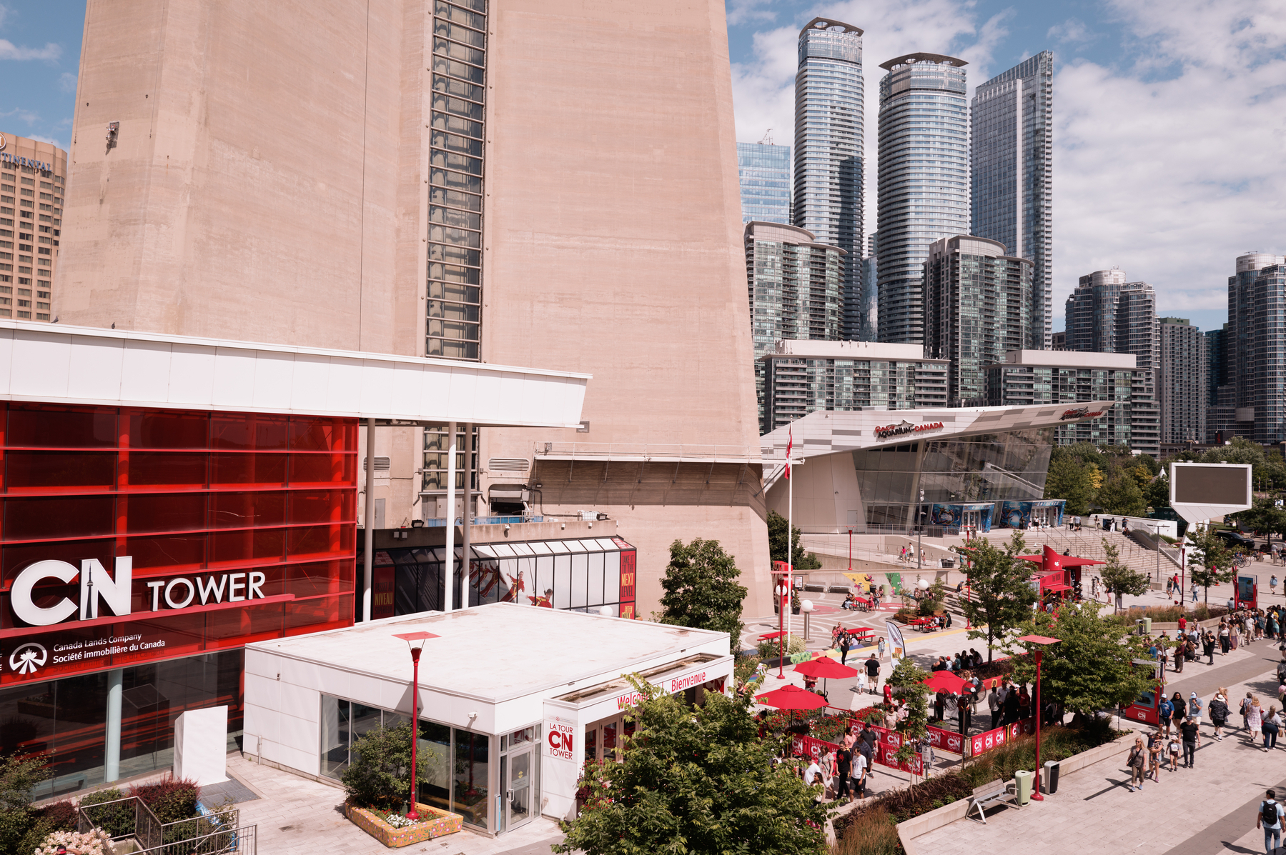 Image showing the entrance to the CN Tower, the base of the CN Tower and in the distance on the right the side of Ripley‘s Aquarium. Lots of people moving around on the square. Image taken from the upper level promenade in front of the Blue Jays Stadium. 