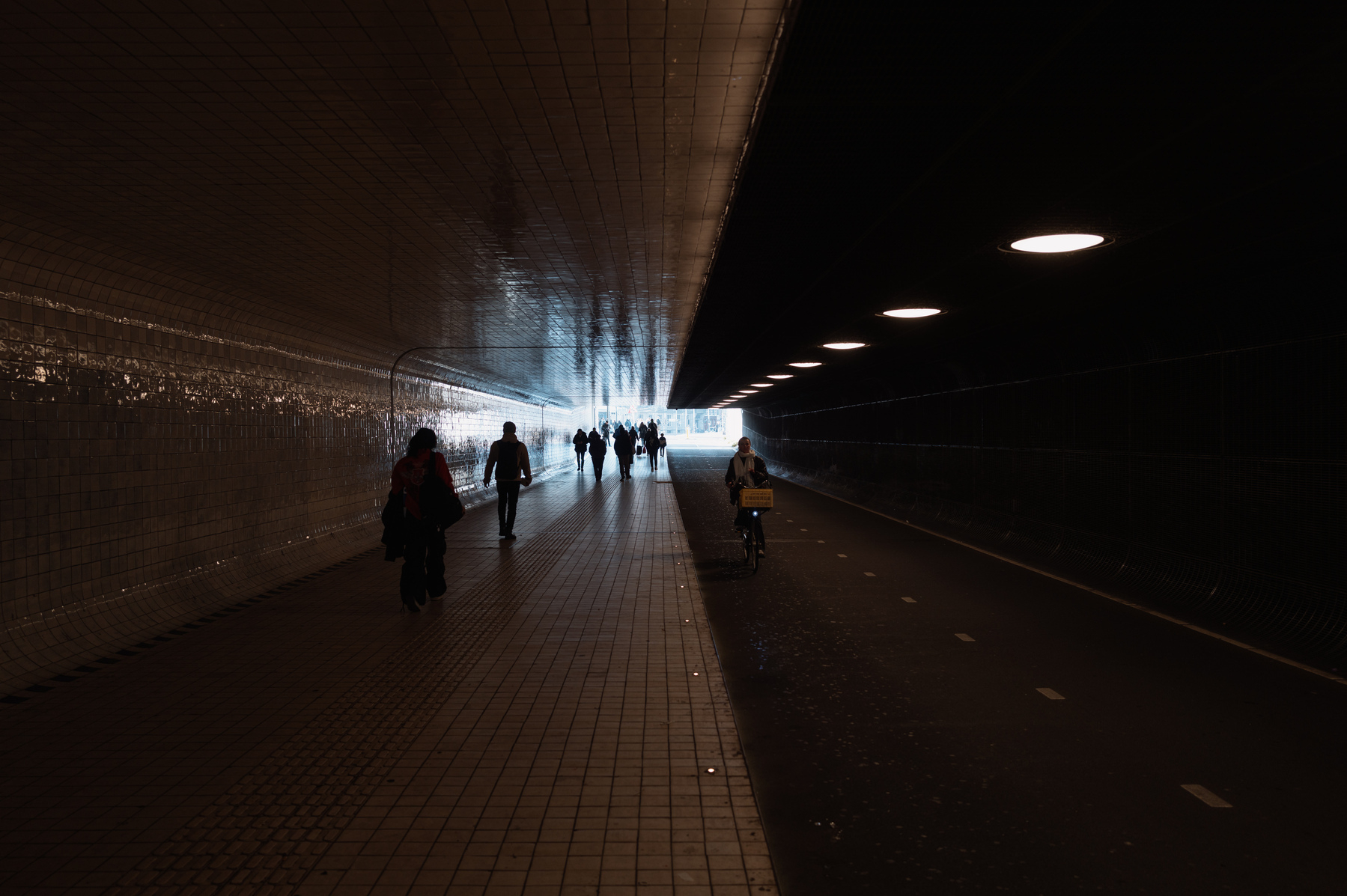 A wide, tiled tunnel at the Amsterdam Centraal station leading towards the city centre. The lights in the tunnel are dim and there is a lot of light streaming in from the other side of the tunnel. On the left a number of people are walking and on the bike lane on the right a person can be seen cycling towards the photographer.