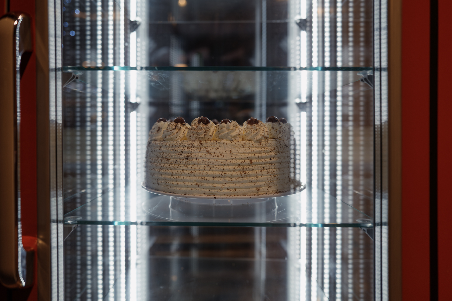 An entire Black Forest gateaux sitting in a glass-doored fridge with glass shelves and lit from the inside. The cake is photographed almost perfectly level from the side. The LED lighting strips on either side of the fridge create vertical reflections that draw the viewer towards the cake sitting in the middle. 