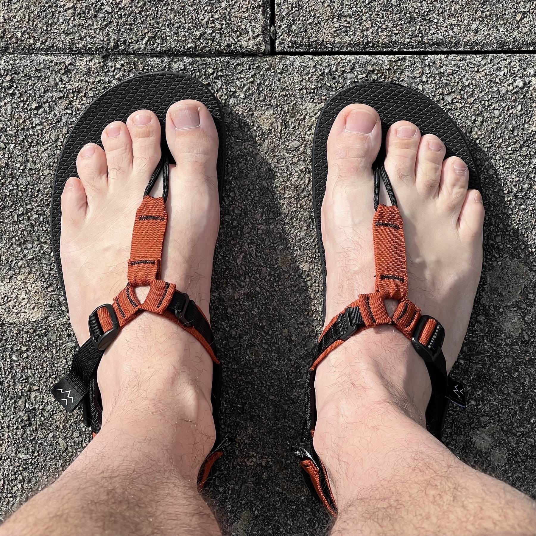 Top down photograph of hairy feet in sandals with a toe separator on top of concrete paving stones. 