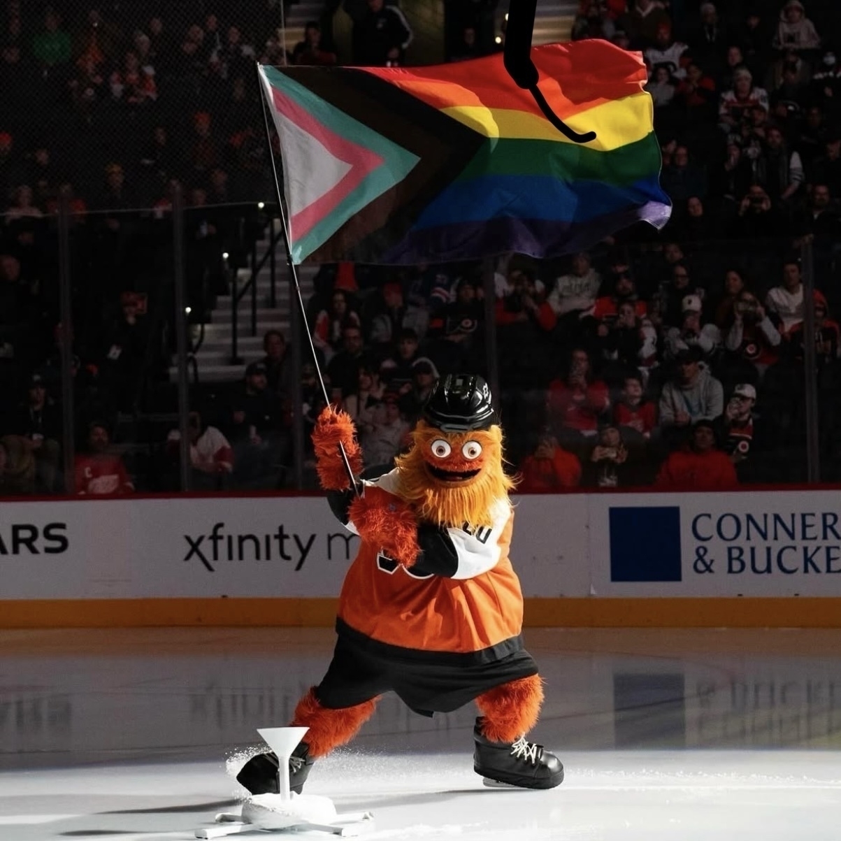 Gritty, the furry orange mascot of the U.S. NHL team „Philadelphia Flyers“ skating in an ice hockey stadium illuminated by a spotlight waving a LGBTQ+ flag.