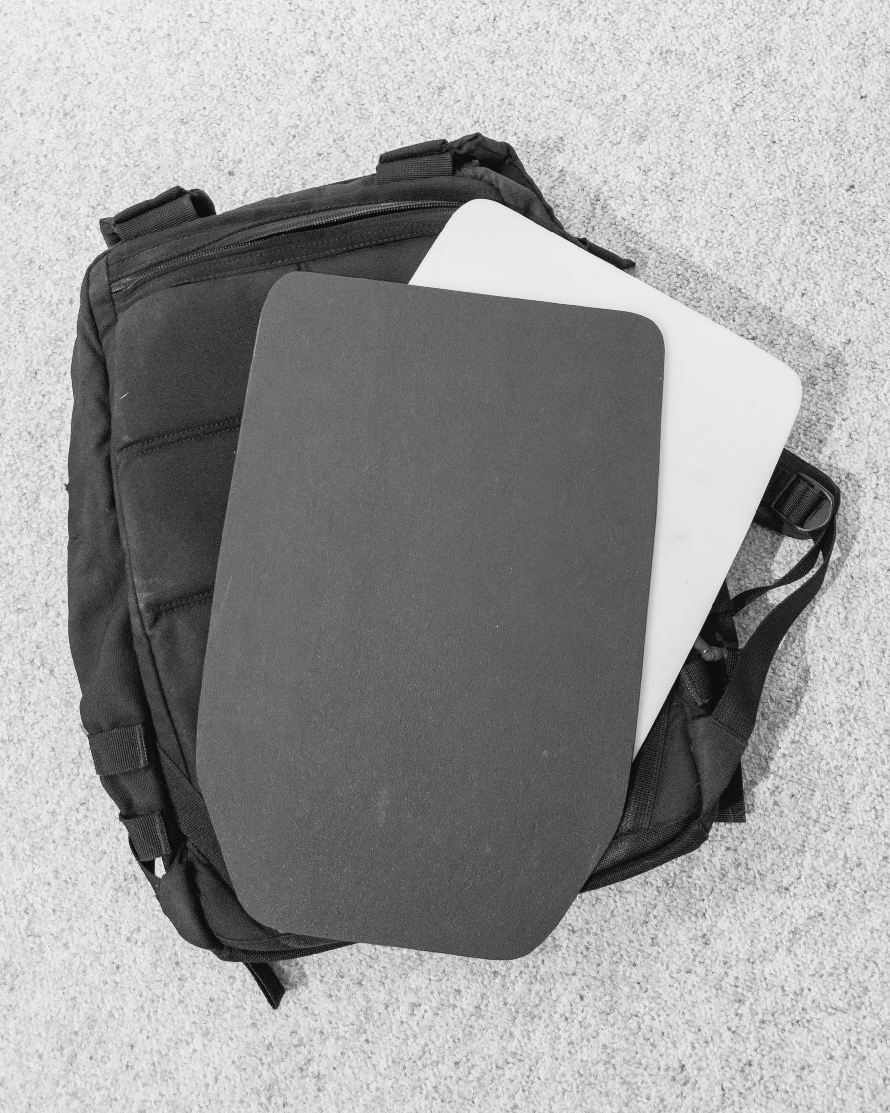 Black and white image of an all-black Goruck backpack laying face down on a carpeted floor. Most of the backside of the pack is obscured by a white, thin frame sheet (a piece of plastic meant to stiffen the backpack and make it more stable and rigid) and a dark grey frame sheet made out of a different material on top of it. The frame sheets are mostly rectangular but have trapezoidal tapers at the bottom.