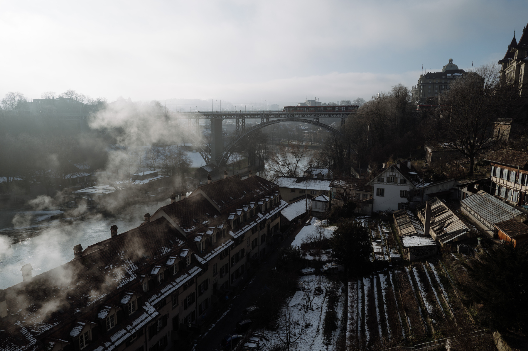 Scene overlooking the river, past worker housing, and rising garden terraces and „Kirchenfeldbrücke“ in Bern from the platform of the Bern munster. The contrast in the image is stark due to harsh light conditions as well as hate and smoke in their air. 