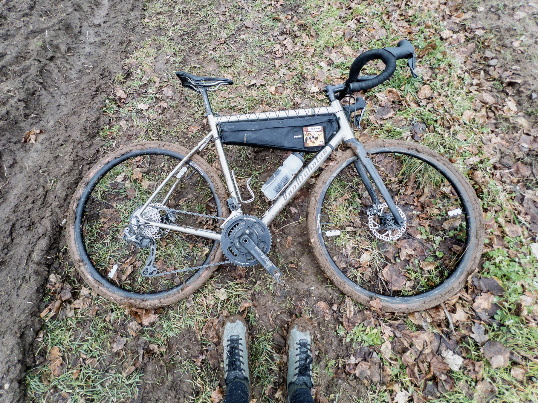 Titanium allroad bike laying on the non-drive side in a muddy field. The perspective has the photographer looking at the bike from below, tires facing the photographer. The tires and rims of the bike are caked in thick, viscous mud. The frame, fork, and full-length frame bag are showing a lot of specks of dirt. Also visible in the image are the feet of the photographer. They‘re wearing blue-green cycling shoes that are dirty and the sides are caked in mud. 