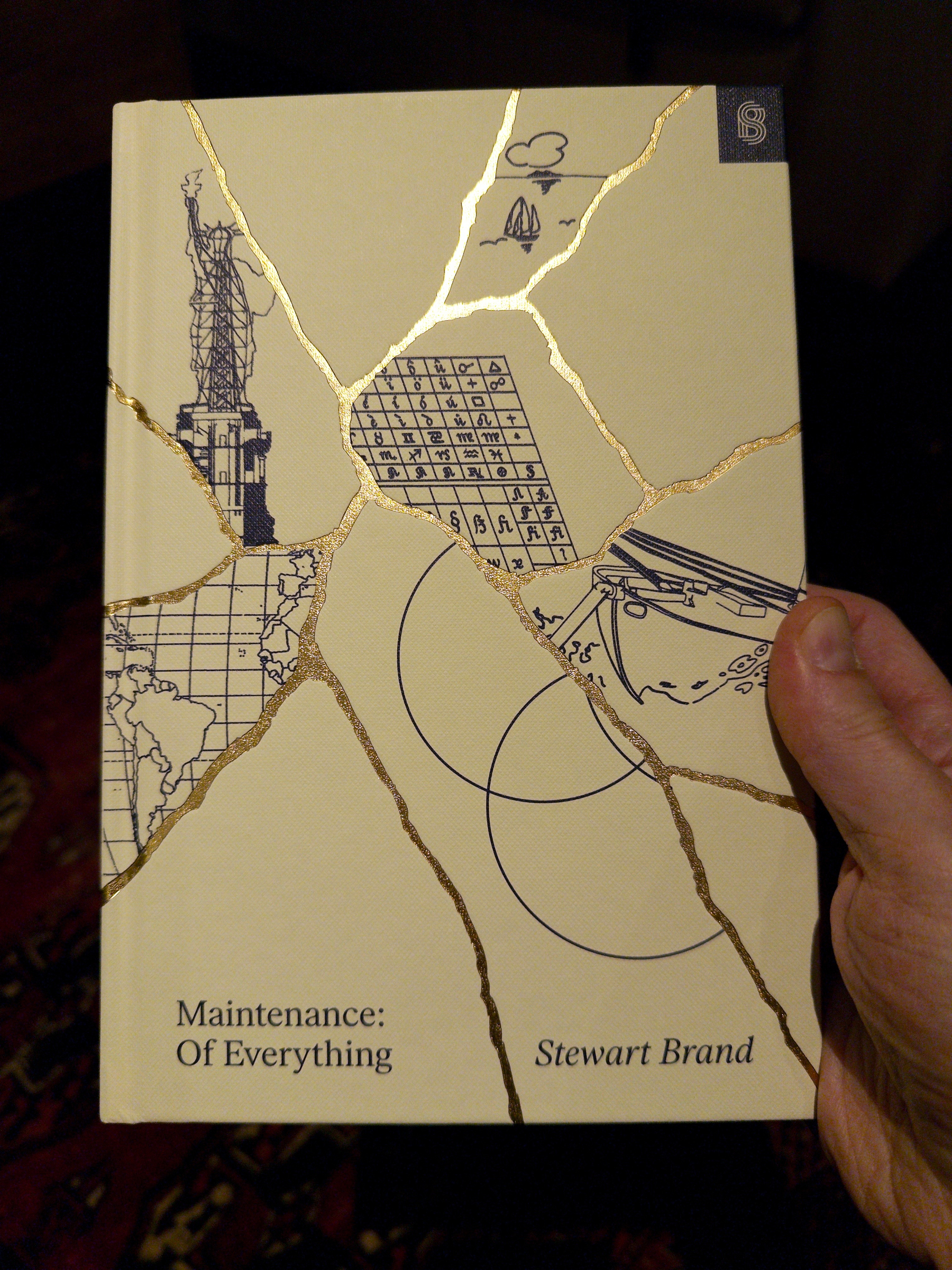 A hand is holding a book titled Maintenance: Of Everything by Stewart Brand, featuring a broken pottery-style design with illustrations on the cover.