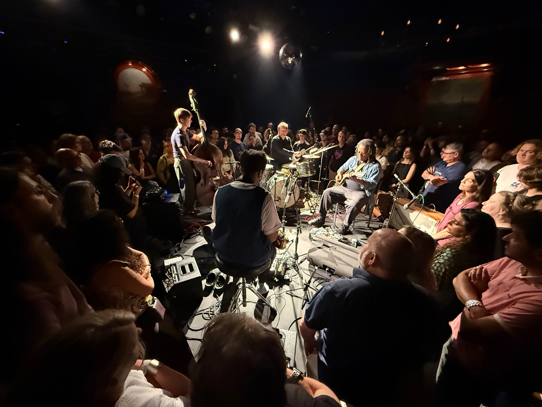 Auto-generated description: A small jazz band is performing live at the center of an intimate, closely-packed audience in a dimly lit venue.