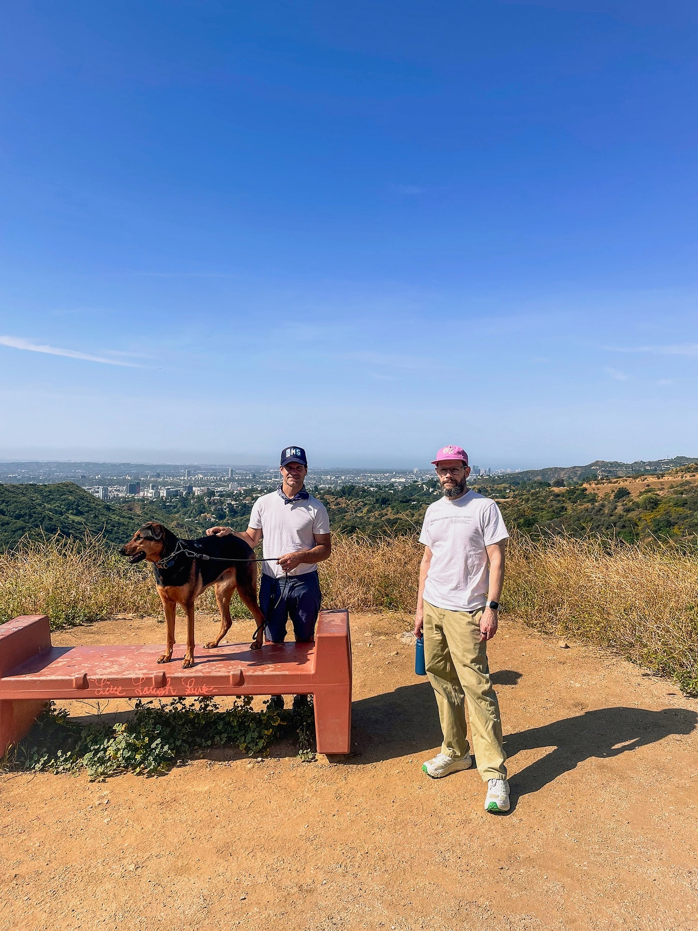 Auto-generated description: Two people and a dog stand on a scenic hilltop with a red bench, overlooking a distant cityscape.