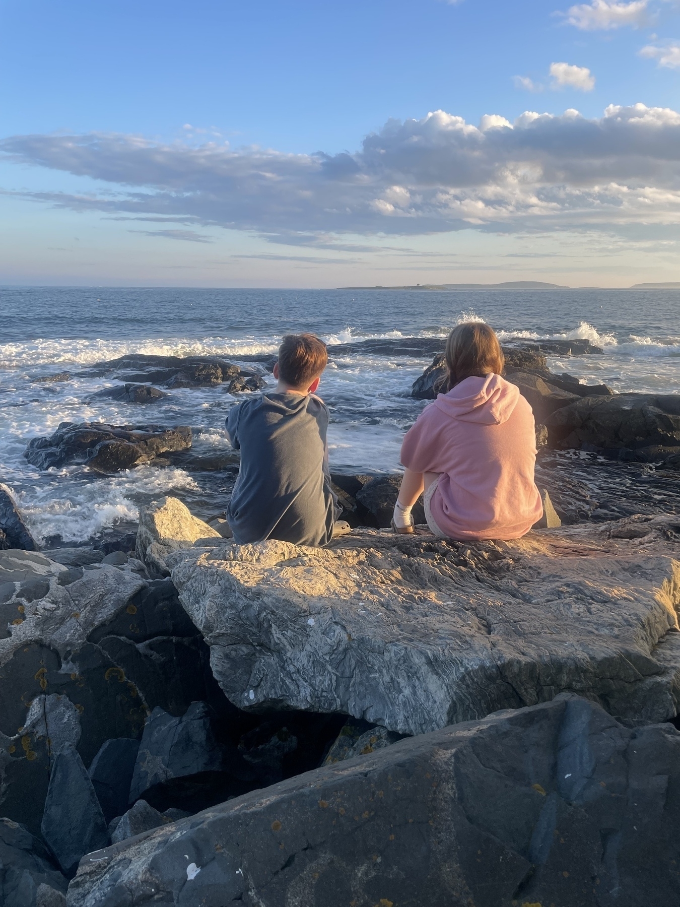 The backs of two teenagers sitting on the rocks on the coast in Maine at sunset 