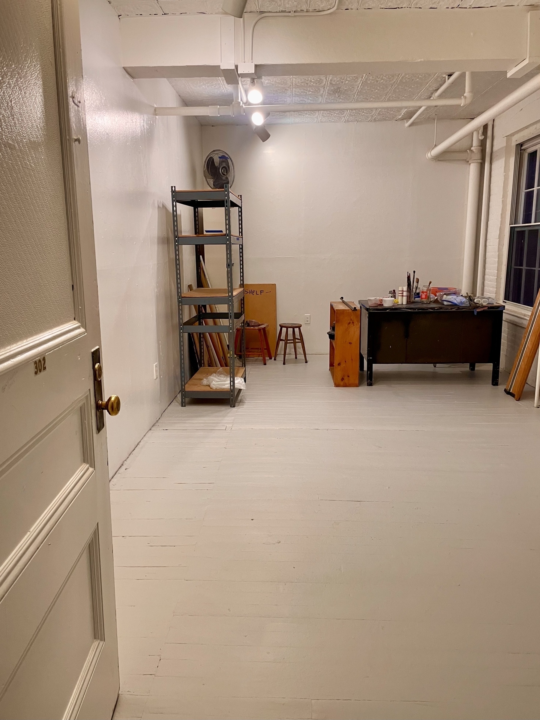 An empty white walled and white floored studio space with desk and shelves, waiting to be populated. 