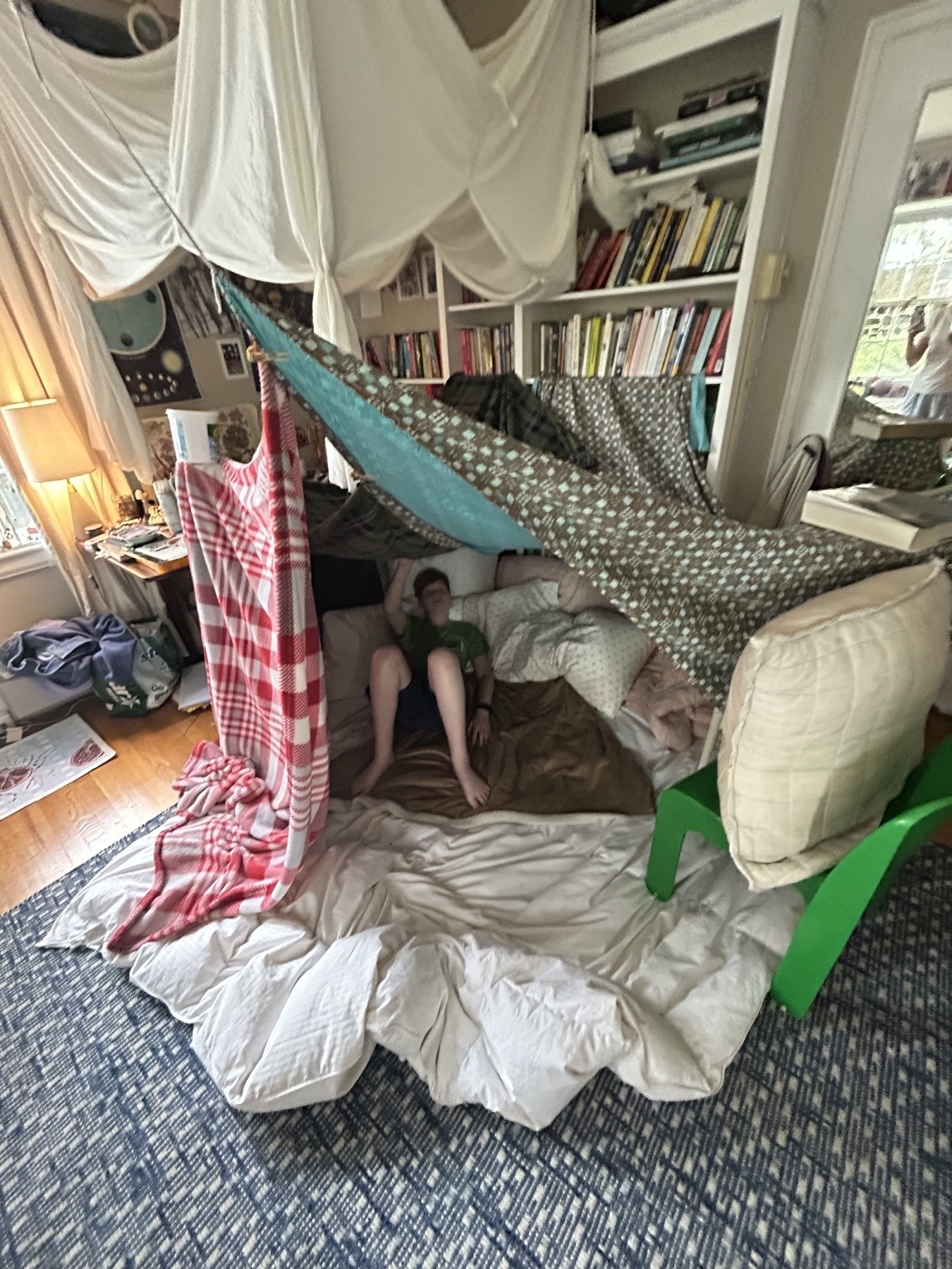 A teenager’s bedroom has an elaborate fort made of blankets and pillows and suspended from chairs and bed frame.