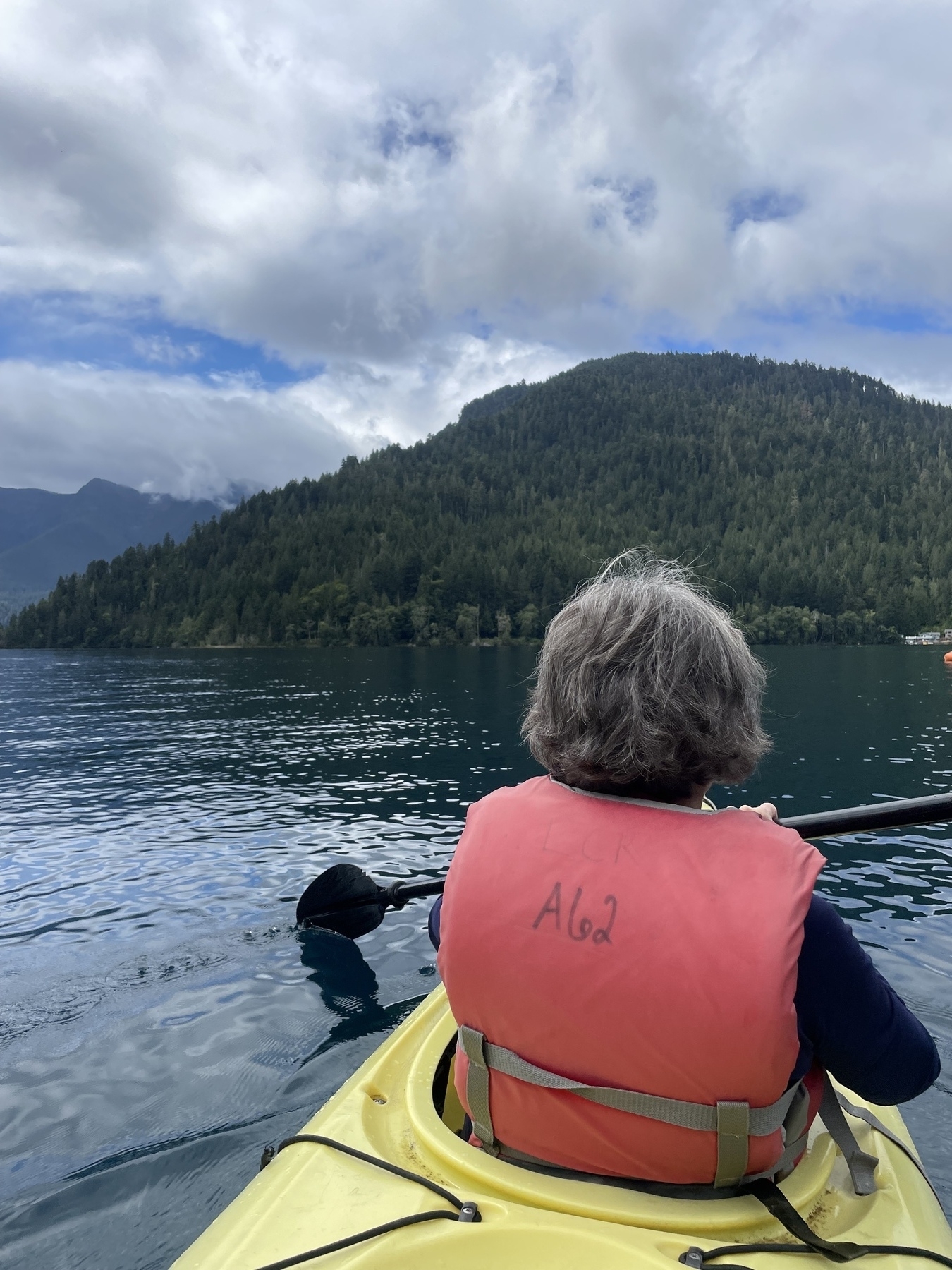 My mom with gray hair on the front of a kayak on Lake Crescent—mountains in the background, blue sky above. 