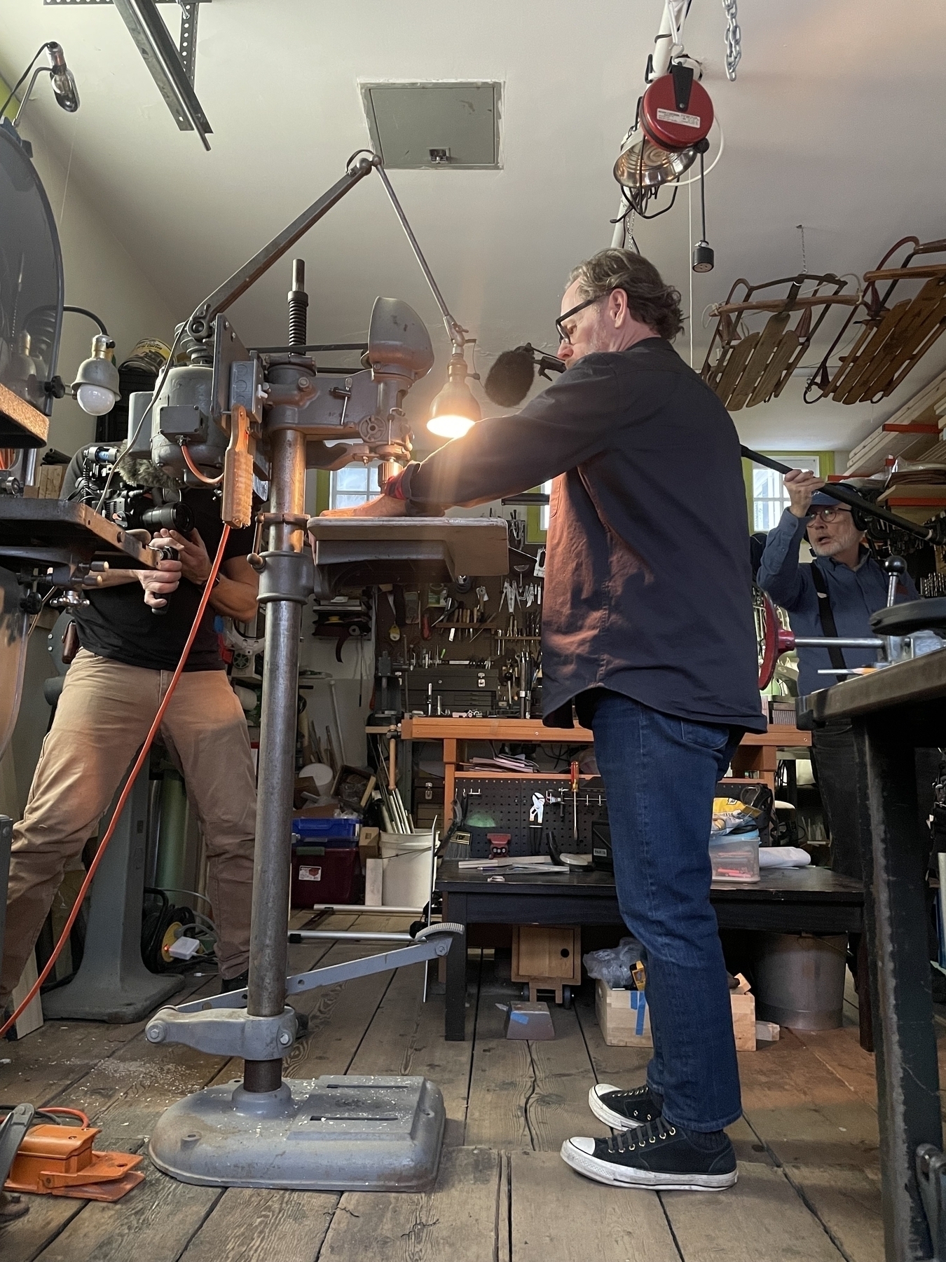 A man with one arm stands at a drill press, while a camera man shoots from left and sound technician holds a microphone at right. 