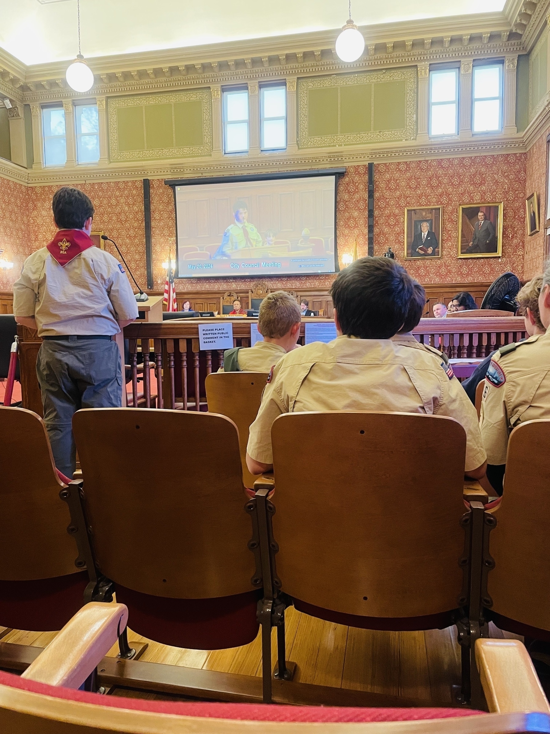 A scout speaks at the mic in a city council meeting, while council members look on.