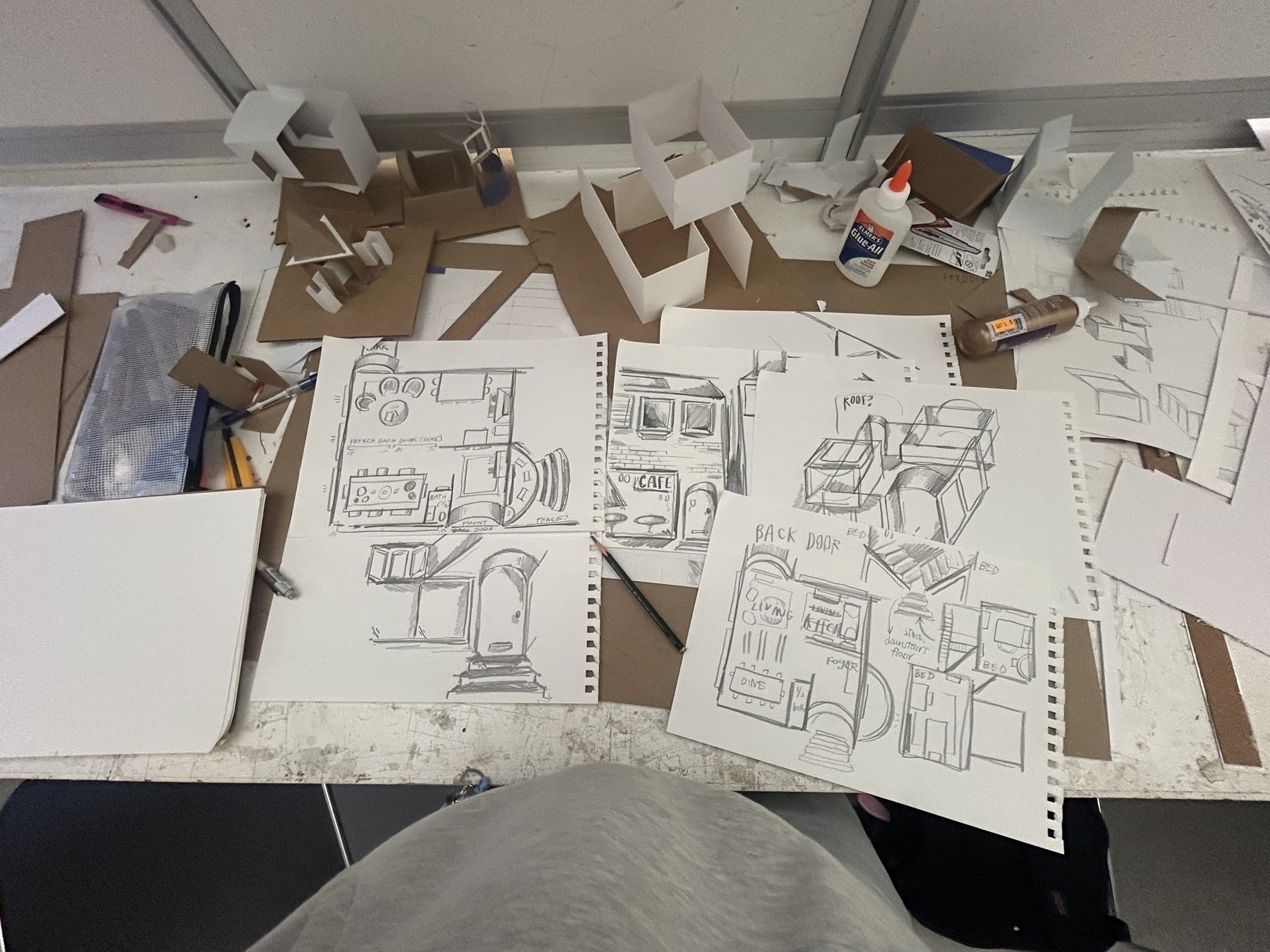 An aerial view of a studio desk has several black and white drawings of house plans and working paper models of buildings
