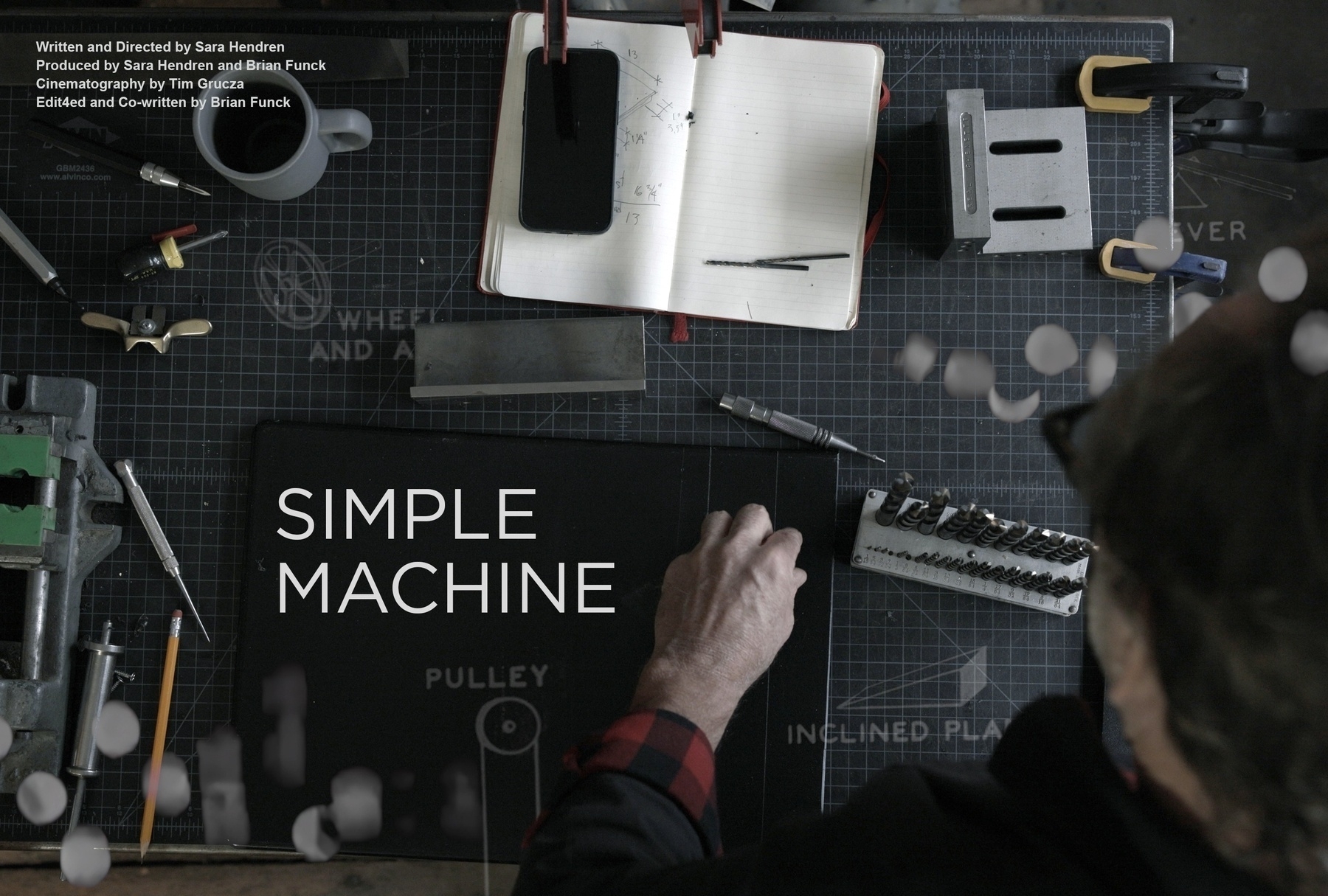 an overhead shot of a work table, with a man's left hand manipulating tools and notebook and more. The words "Simple Machine" are in the middle, with credits on the upper left corner.