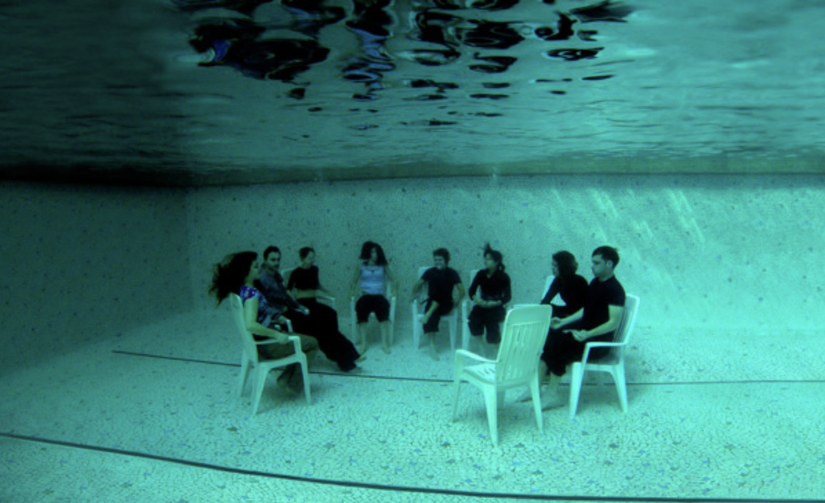 A group of people sits at the bottom of a swimming pool in white plastic chairs, arranged in a circle as for a meeting.