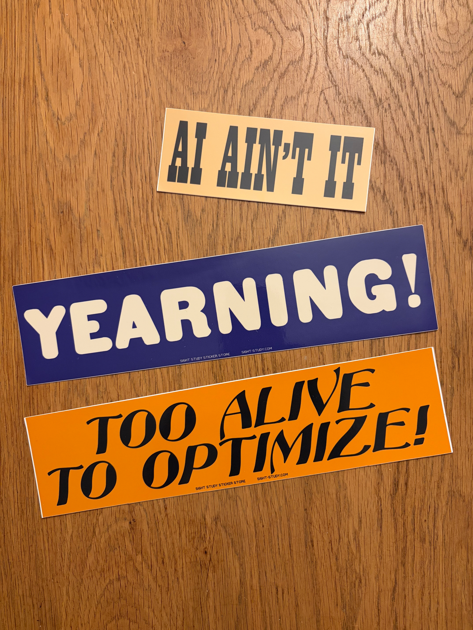 Three bumper-style stickers: one reads "AI Ain't It," one reads "Too Alive to Optimize" and one reads, simply, "Yearning!"