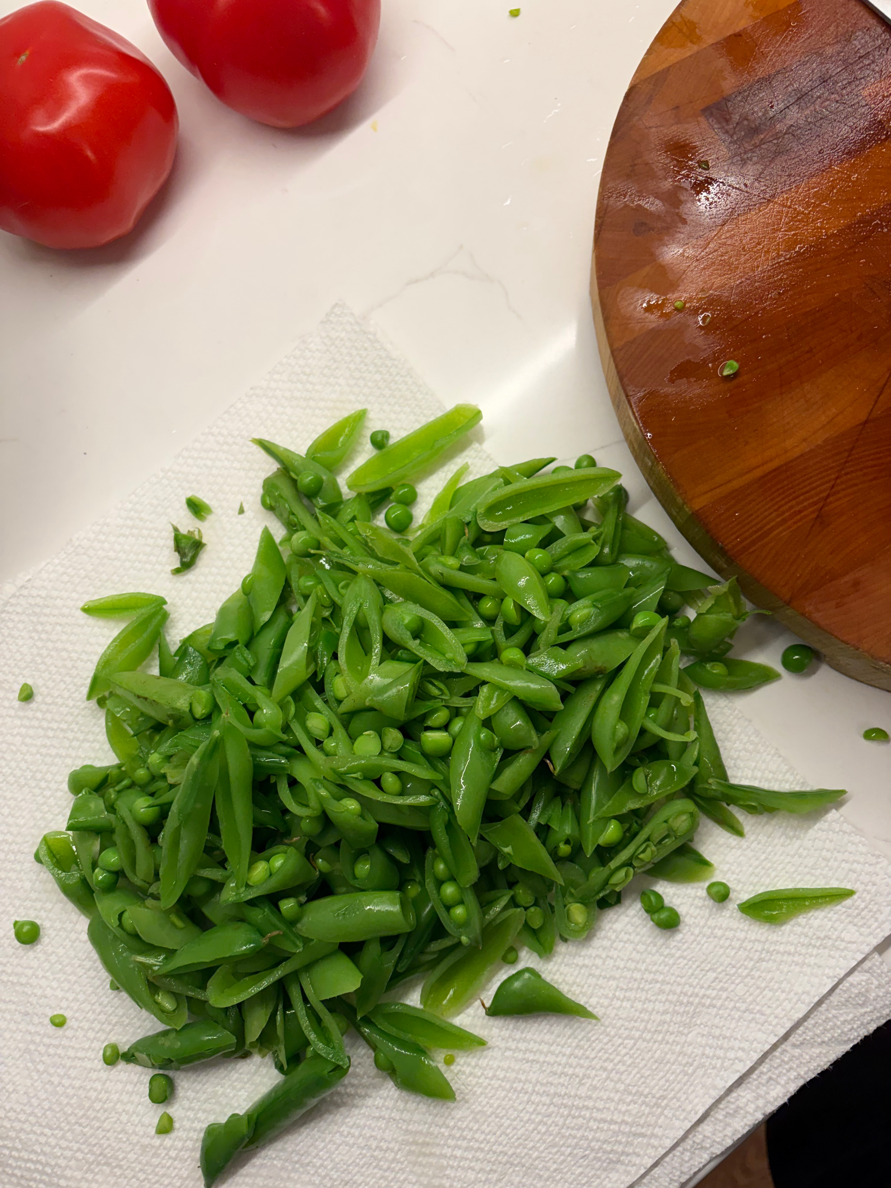 On my kitchen counter, a bright green array of sugar snap peas cut on the bias, with my wooden cutting board and a couple of tomatoes nearby. 