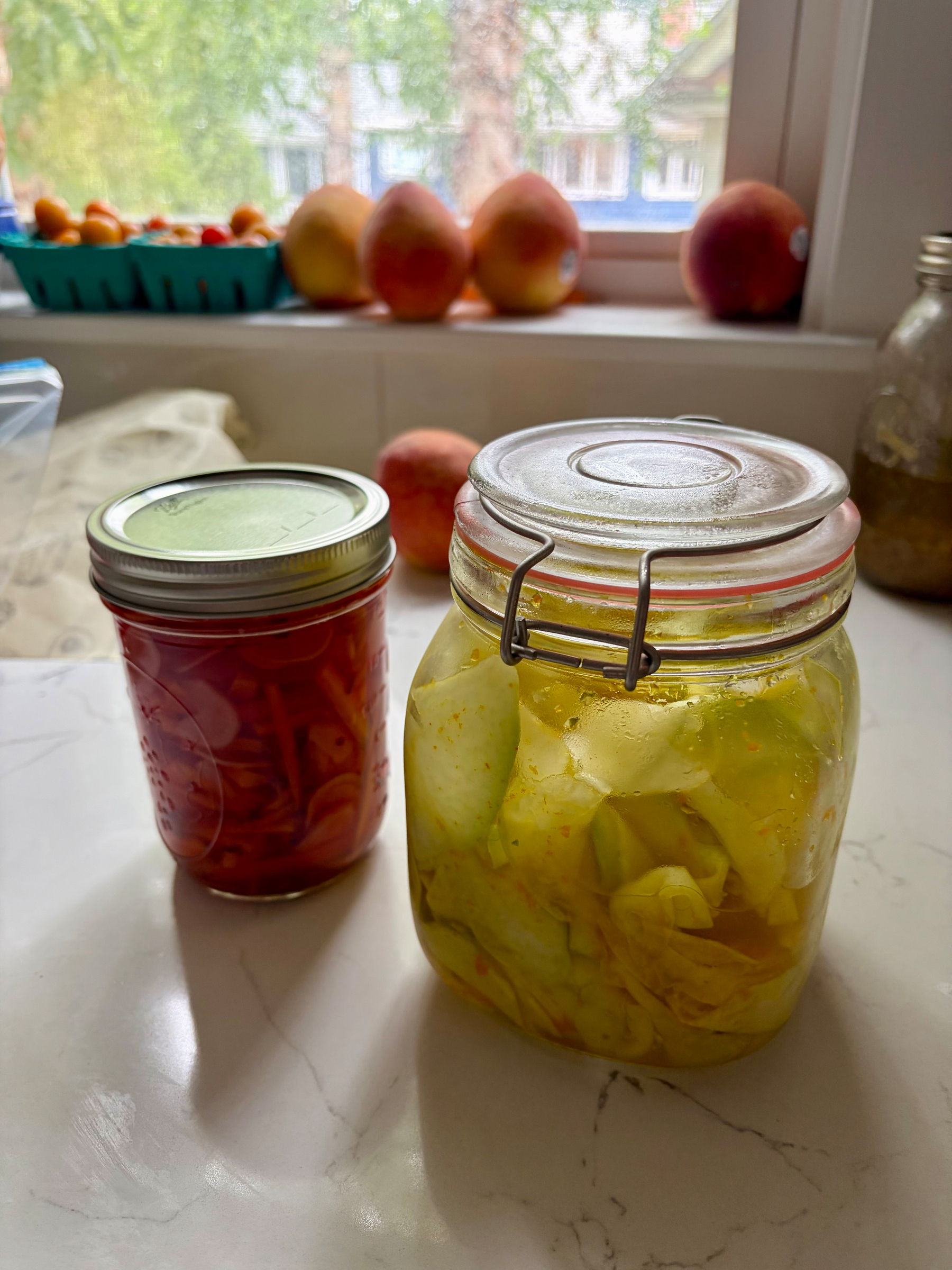 On my kitchen counter, two jars of colorful summer pickles, with fresh peaches and cherry tomatoes lining the window sill in the background 