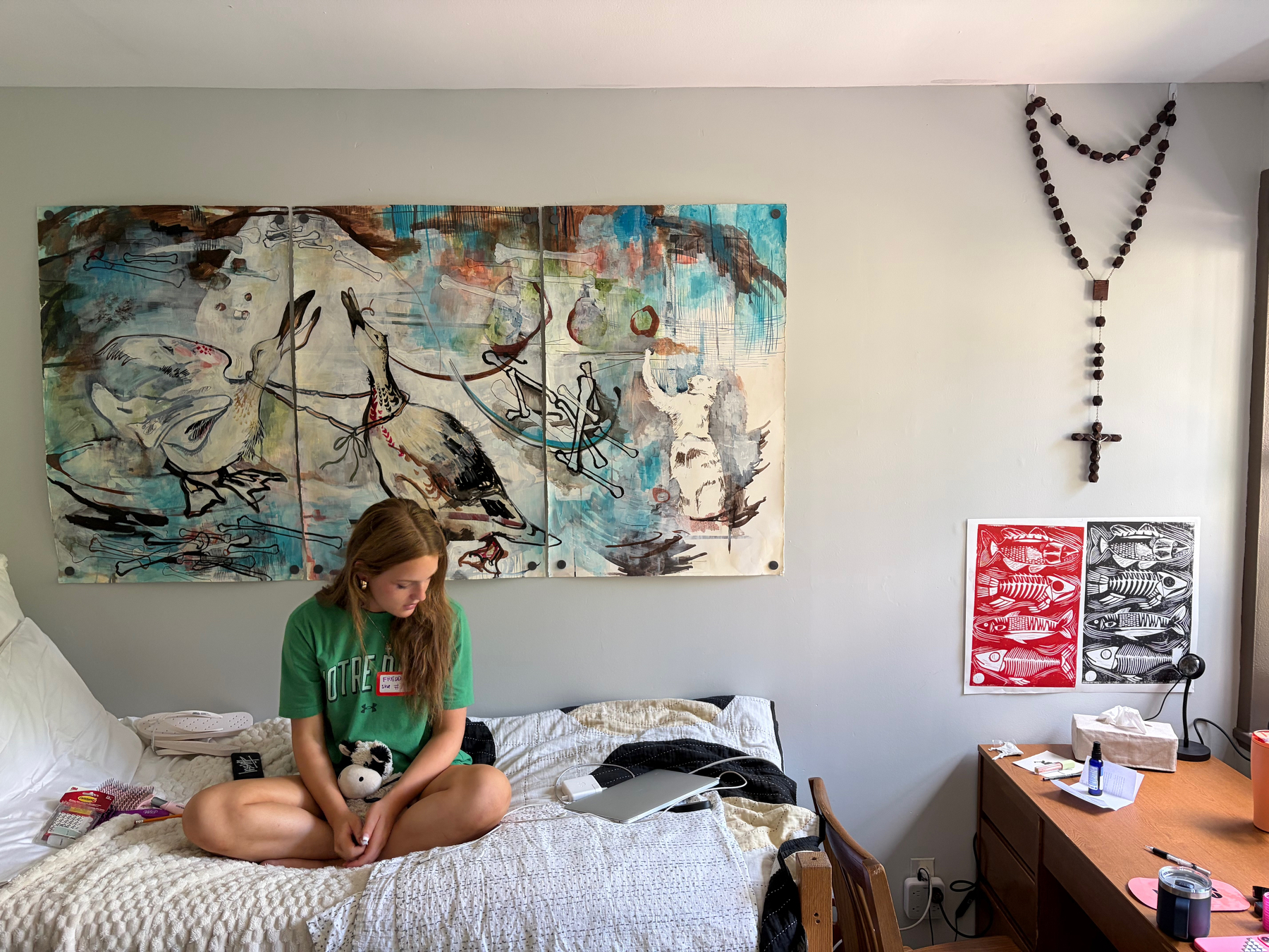 Our daughter sits on her dorm room bed with a large triptych painting behind her, two sets of lino prints featuring rows of fish, and a large wooden rosary on the wall.