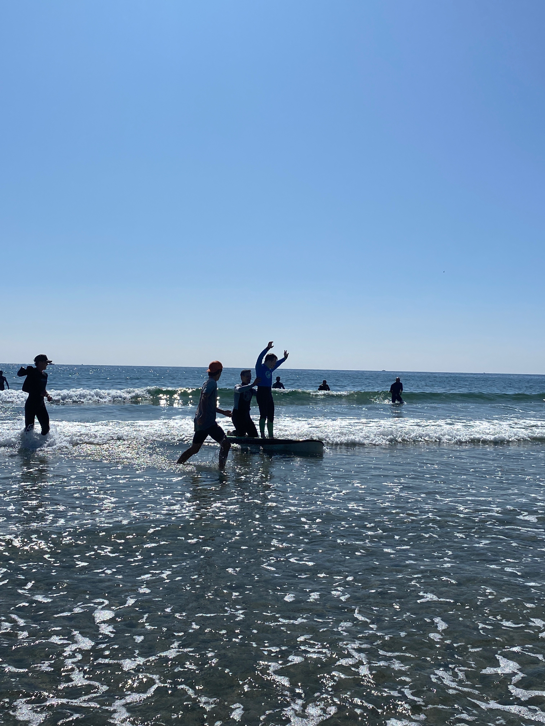 Graham, both arms raised, coasts into the shoreline, standing on a surfboard and supported from behind. Other surfers chase alongside him. 