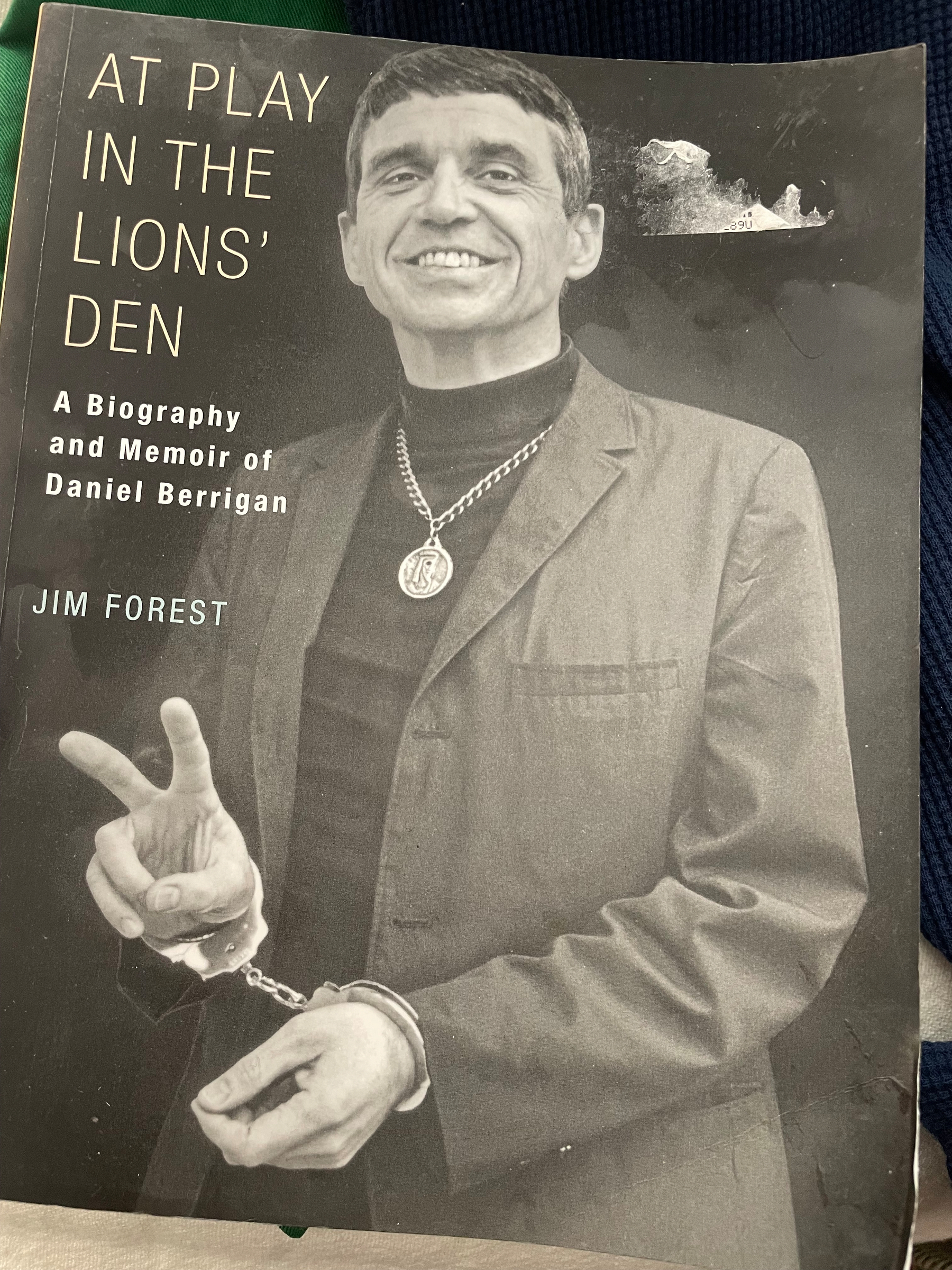 The cover of Jim Forest’s At Play in the Lion’s Den. Fr Berrigan stands in handcuffs and making a peace sign. 
