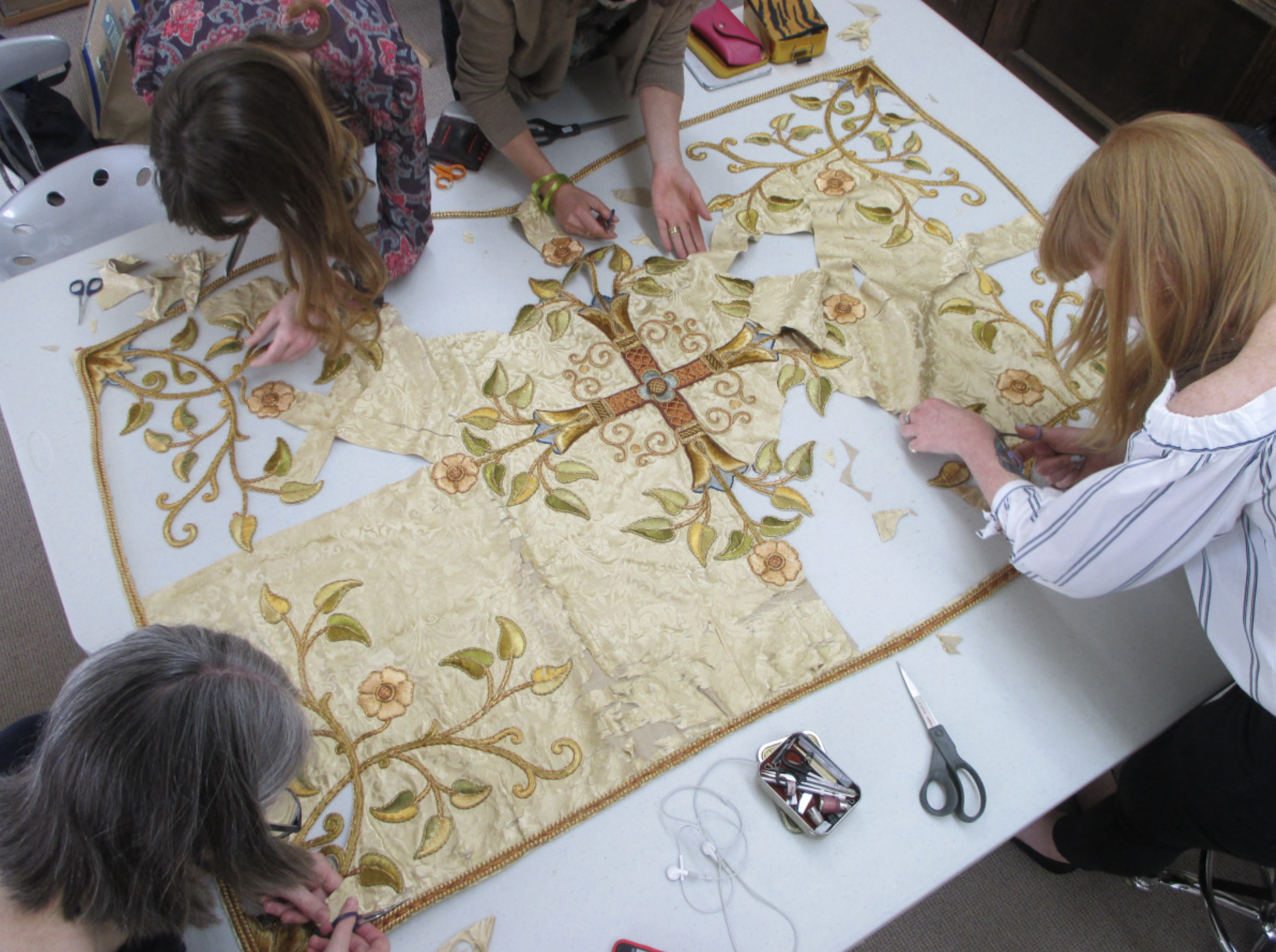 An overhead shot of an elaborate silk reredos, with four conservators working around it with needles and scissors. It's got a cream background, cross in the middle, and green and red and orange floral patterns on it.