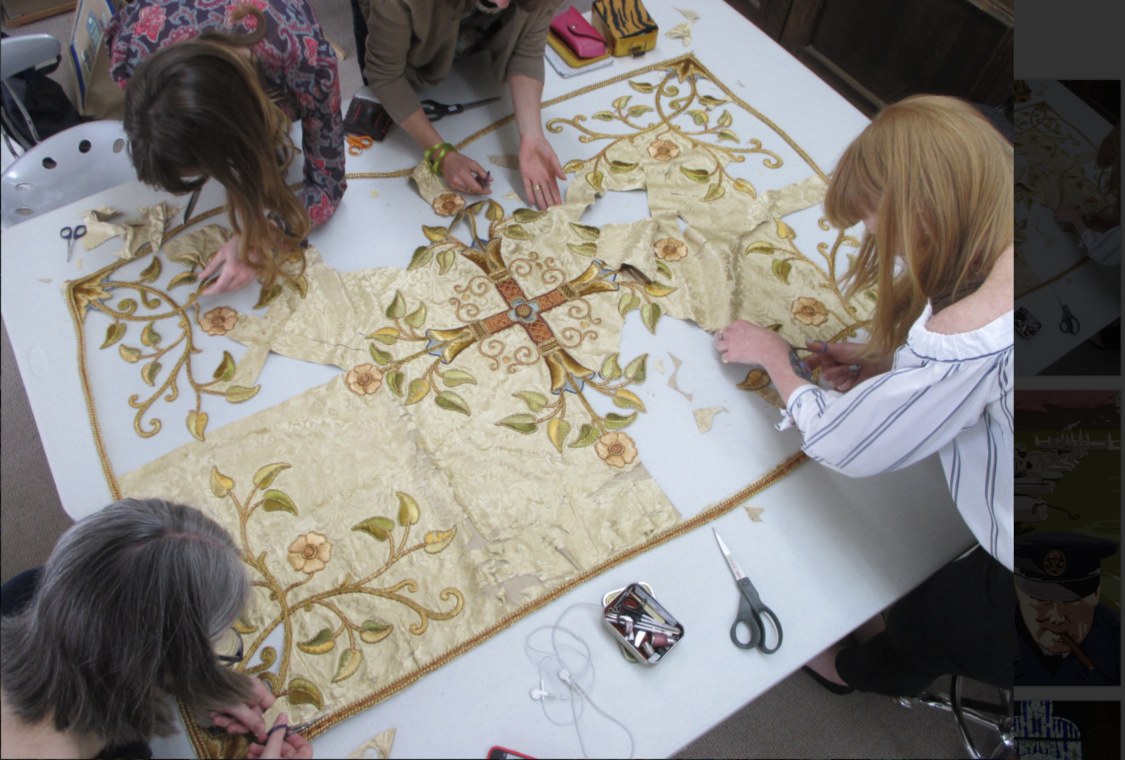 An overhead shot of a table with an ornate silk tapestry, of cream and red and green, with a cross in the center and ornamentation around the edges. Four women work carefully on various parts to mend it.