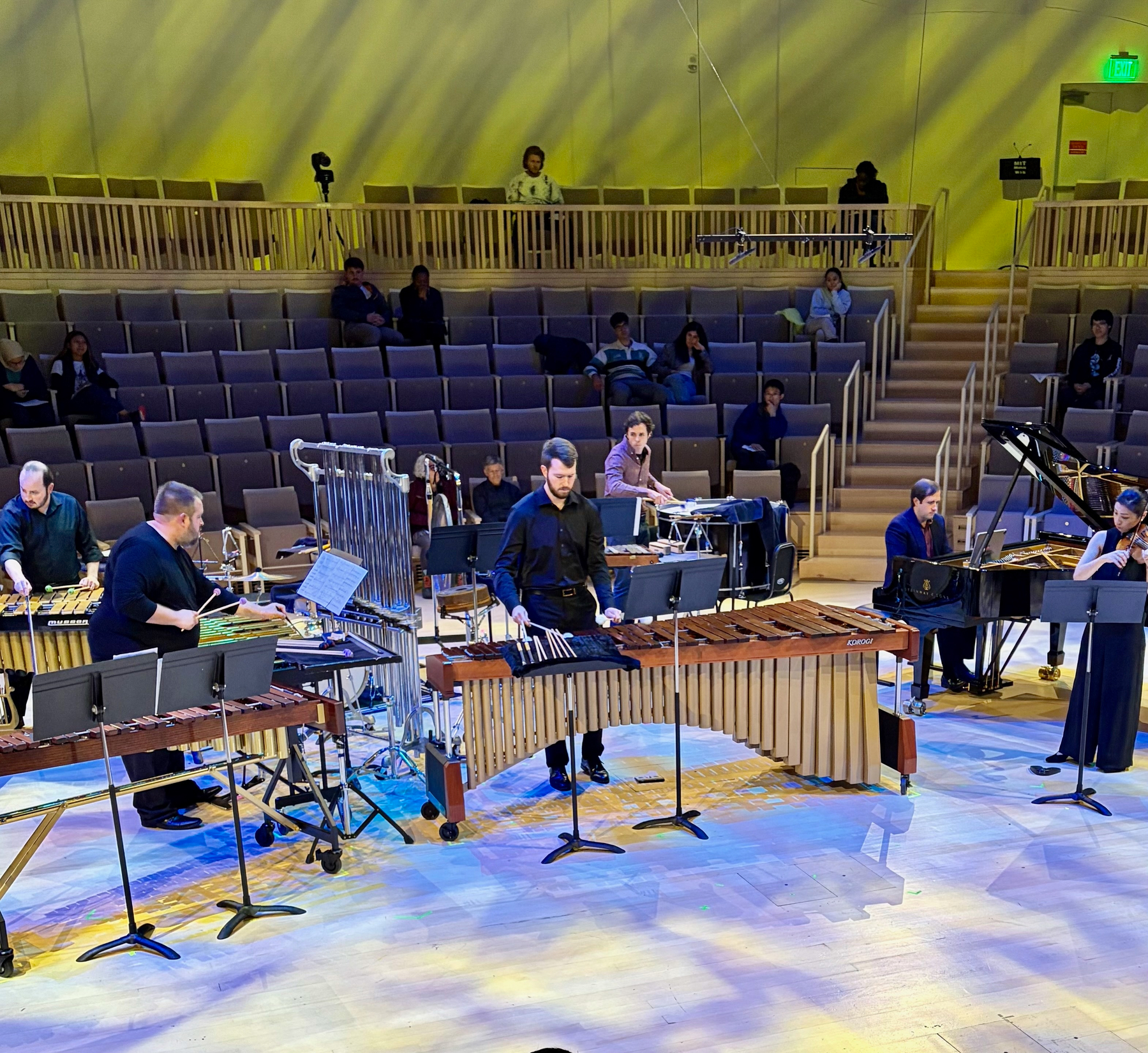 In MIT's in-the-round Thomas Tull concert hall, half a dozen musicians play timpani, drums, gongs, and other percussion instruments, while another plays piano and another violin. Yellow and blue light pervade the scene.