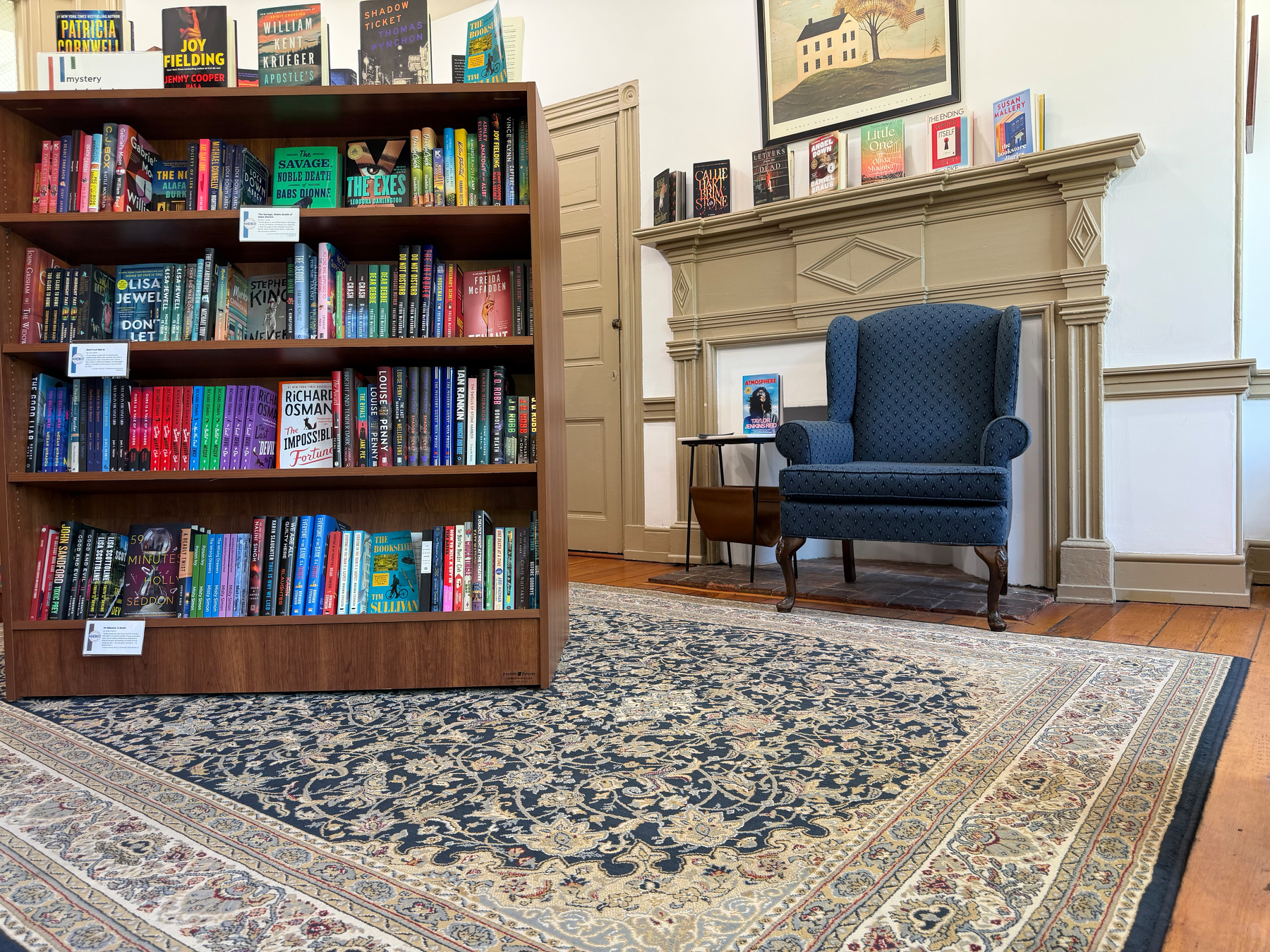 A cozy reading corner features a bookshelf filled with colorful books, a blue armchair, and a decorative rug.