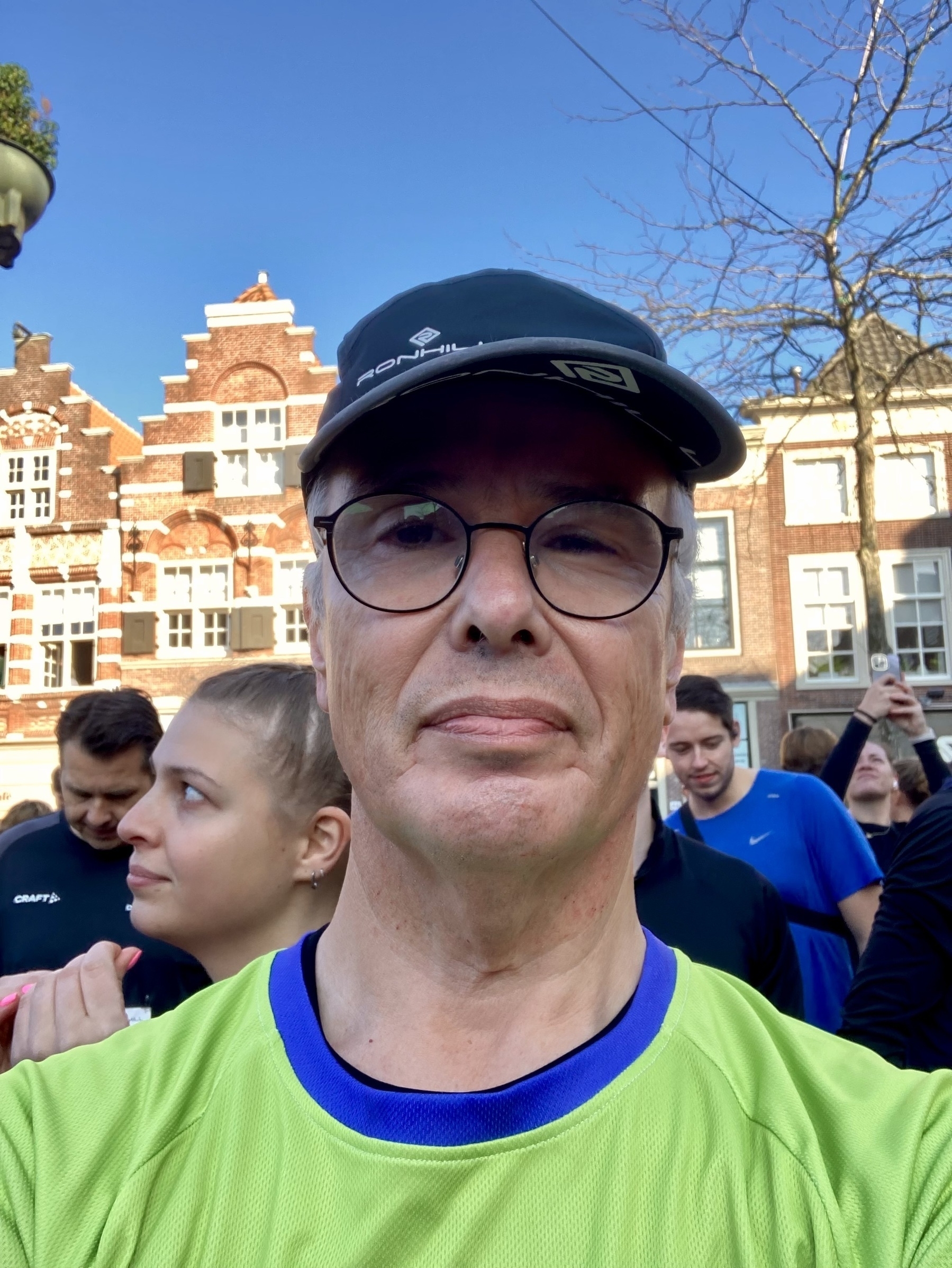 Een persoon met een pet en bril maakt een selfie in een groepsomgeving met gebouwen op de achtergrond.