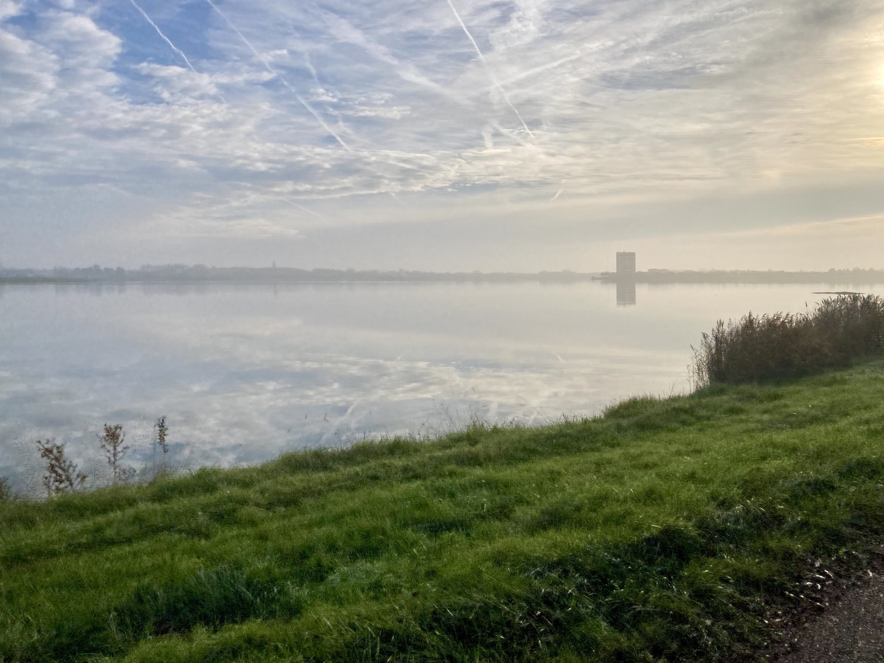 Een serene uitzicht op het meer met een met gras begroeide voorgrond, kalm water dat de bewolkte lucht weerspiegelt en een ver silhouet van een gebouw.