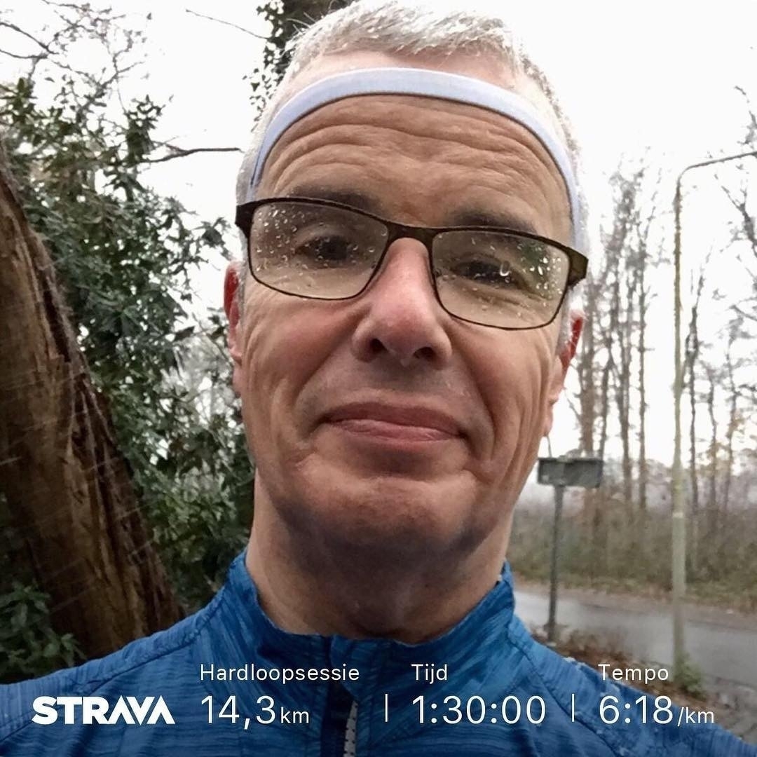 Een man met een bril en een witte hoofdband maakt een selfie buiten op een pad in een bosrijk gebied, met Strava-oefeningsstatistieken die de afbeelding overlappen die wijzen op een run van 14,3 km in 1 uur en 30 minuten in een tempo van 6:18 per km.