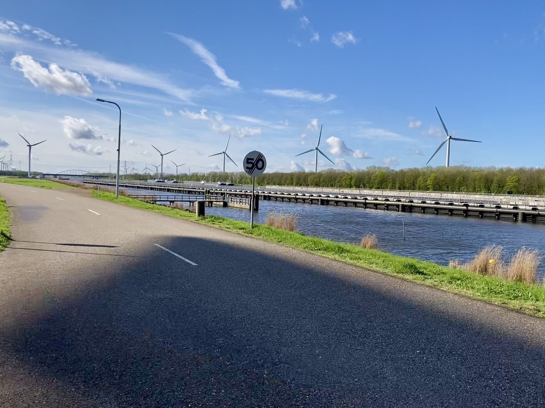 Auto-generated description: Een verharde weg loopt langs een kanaal, met een snelheidslimietbord van 50 km/u en windturbines op de achtergrond onder een gedeeltelijk bewolkte hemel.