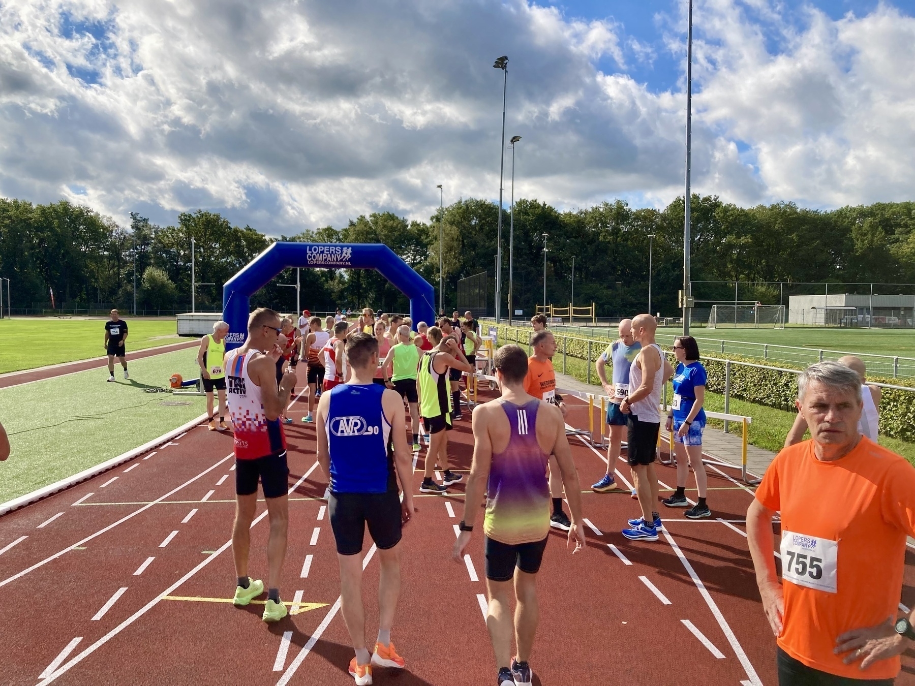 Hardlopers verzamelen zich bij de startlijn van een buitenbaanevenement onder een blauwe lucht met verspreide wolken.