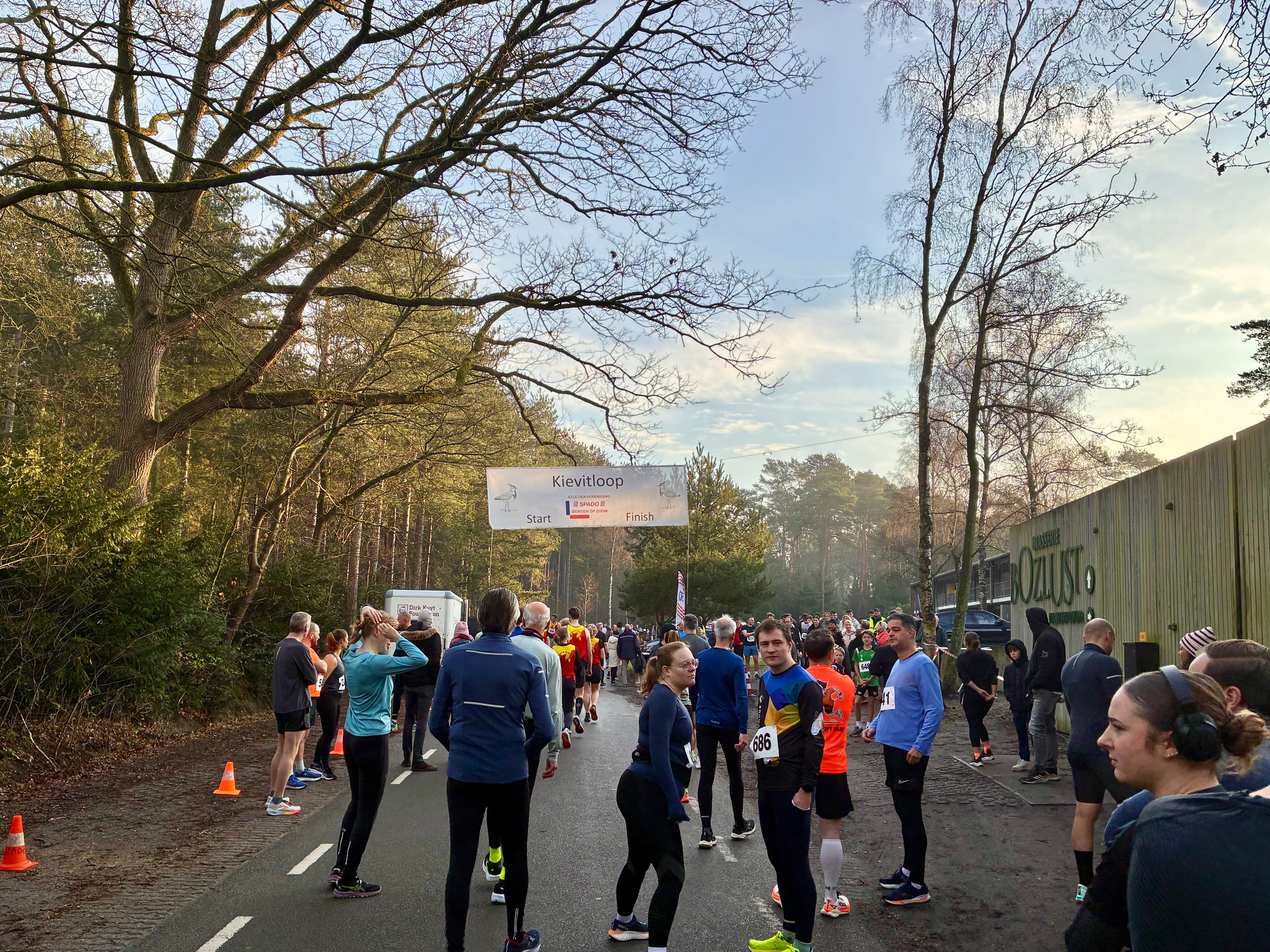 Een groep hardlopers is verzameld op een weg in de buurt van een park, om zich voor te bereiden op of deel te nemen aan een wedstrijd.