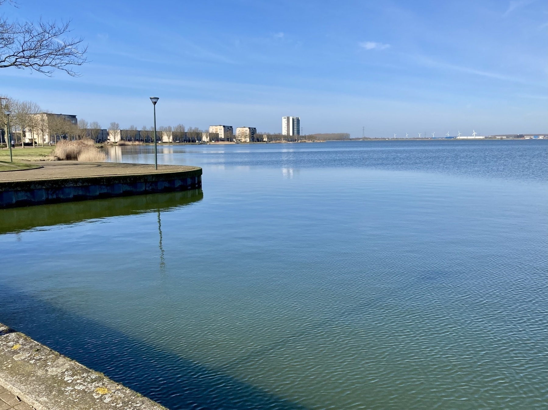 Een rustig waterlichaam reflecteert de helderblauwe lucht, begrensd door een kade en verre gebouwen.