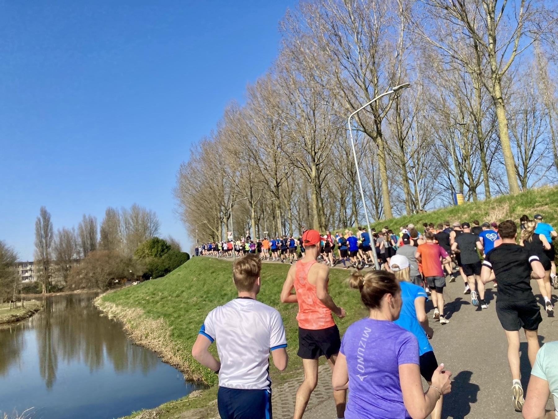 Een groep mensen rent op een zonnige dag langs een pad naast een kanaal, omringd door bomen.