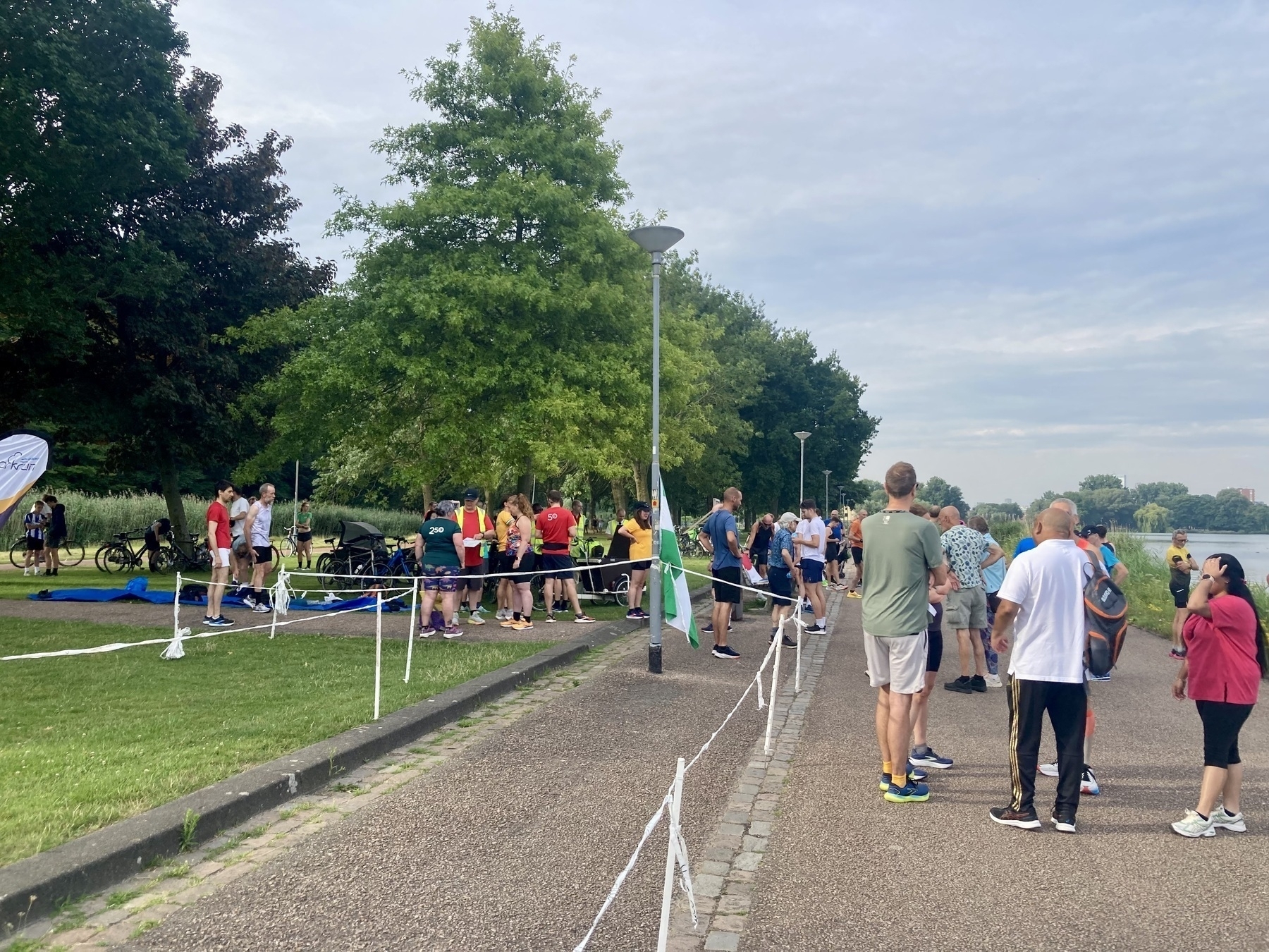 Een groep mensen verzamelt zich in de buurt van een pad, sommigen in sportkleding, terwijl ze deelnemen aan een buitenevenement naast een grasveld en bomen.