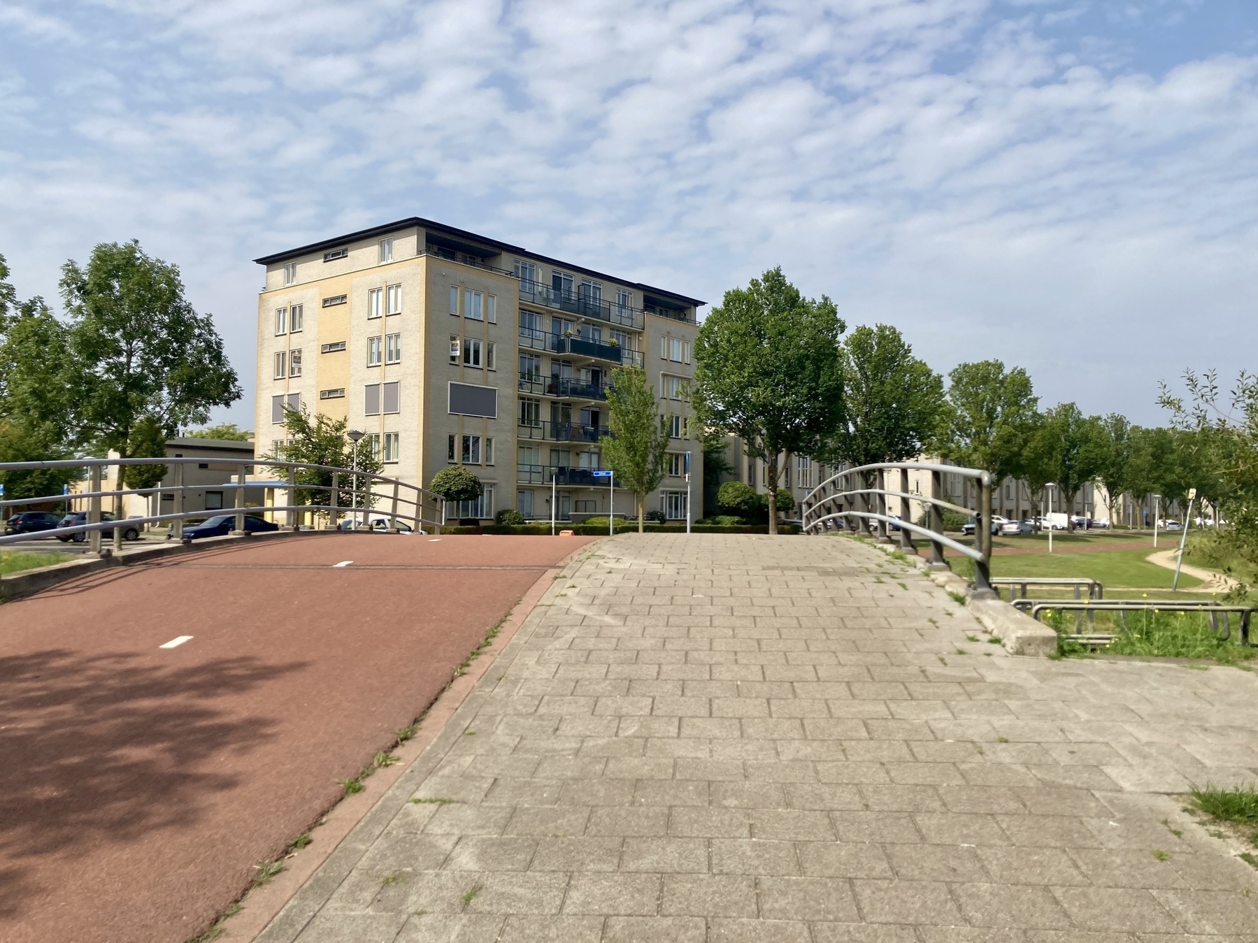 Een verhard pad leidt over een kleine brug, geflankeerd door een woongebouw en groene bomen onder een deels bewolkte lucht.